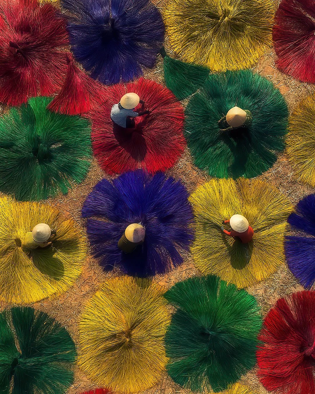 People with traditional conical hats walking among large, colorful, umbrella-shaped straw decorations in a sandy area.