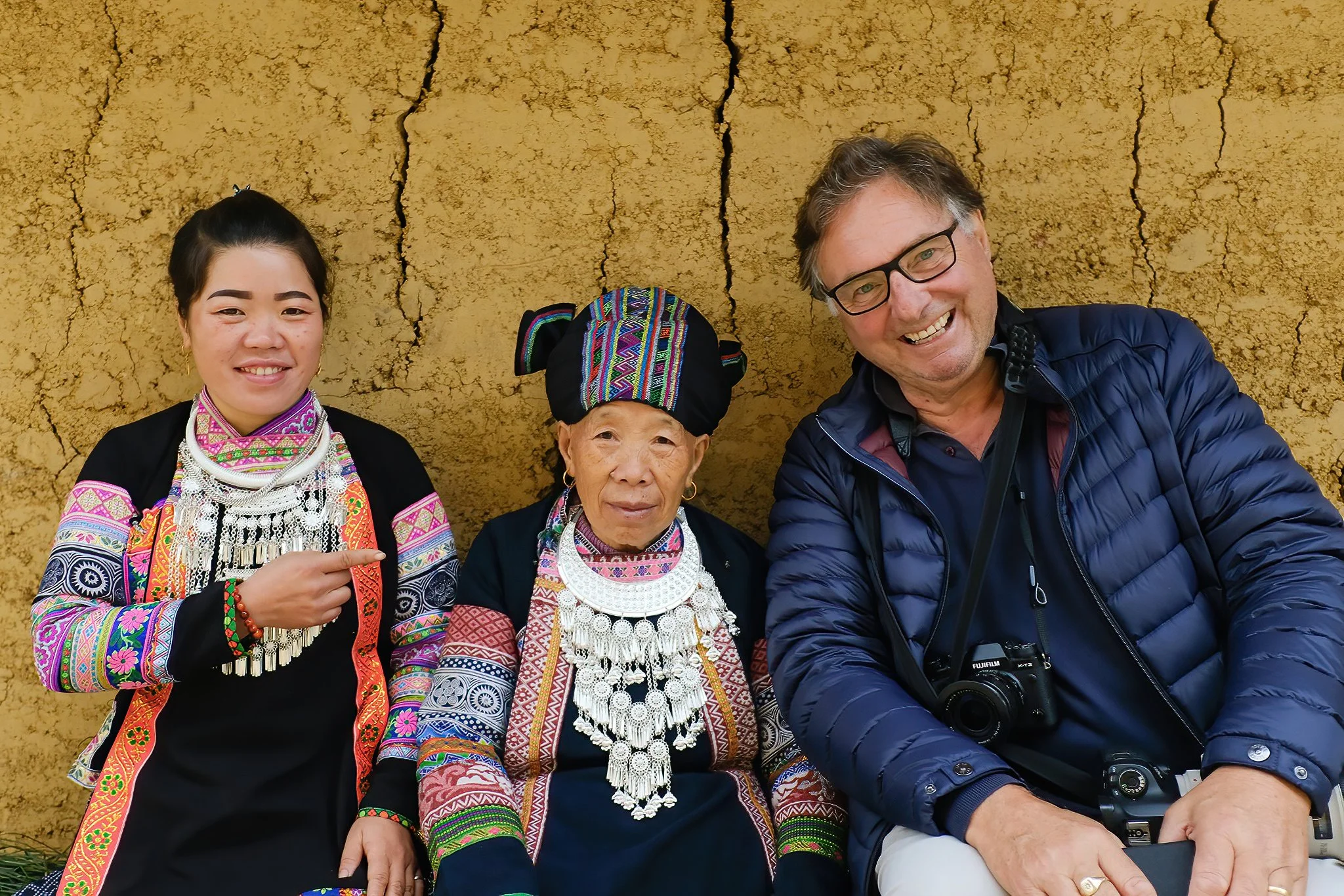 Three people sitting in front of a textured, tan-colored wall. The person on the left is a young woman with black hair in a bun, wearing traditional colorful clothing and jewelry, and pointing at the middle person. The middle person is an older woman with gray hair under a black headwrap, also dressed in traditional clothing and jewelry. The person on the right is a middle-aged man with glasses, wearing a navy blue jacket, holding a camera, and smiling.