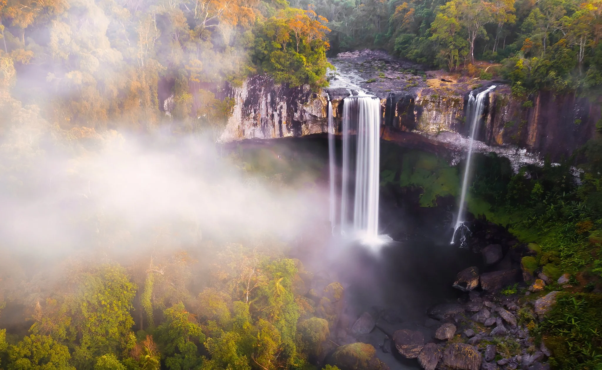 A scenic view of a waterfall cascading down a cliff into a river, surrounded by lush green trees and mist in the air.