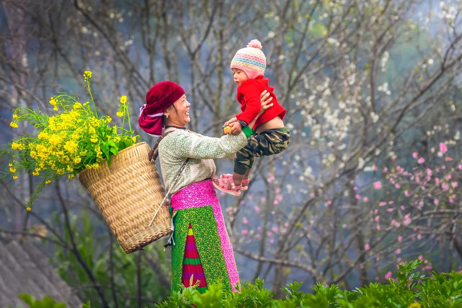 A woman in colorful traditional clothing carrying a basket of yellow flowers on her back, smiling and holding a young child in a red sweater with a pink, white, and blue knit hat, outdoors with blooming trees.