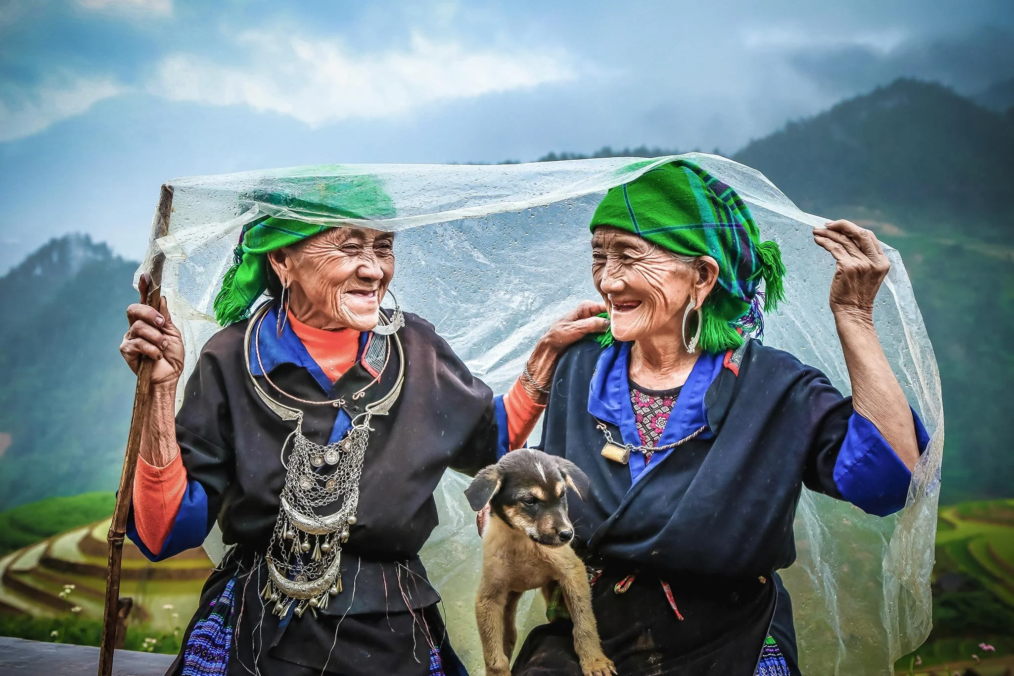 Two elderly women wearing green plaid headscarves and colorful jewelry smile at each other under a clear plastic sheet, with a rural hillside landscape in the background. A puppy hangs from one woman's arm.