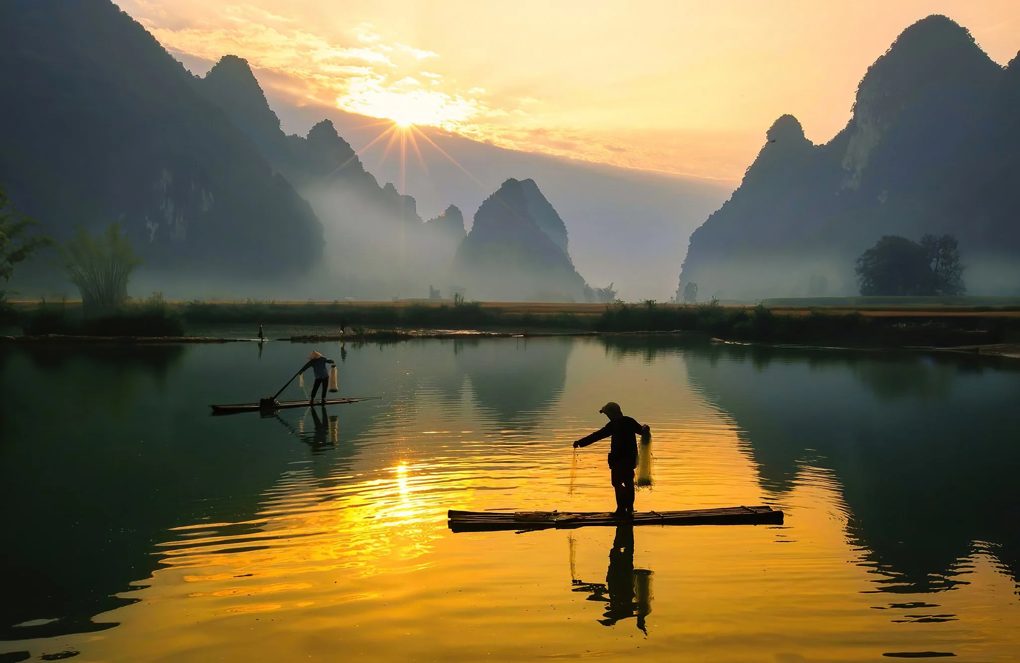 Silhouettes of two people on bamboo rafts in a lake at sunset, surrounded by mountainous landscape with mist and colorful sky.