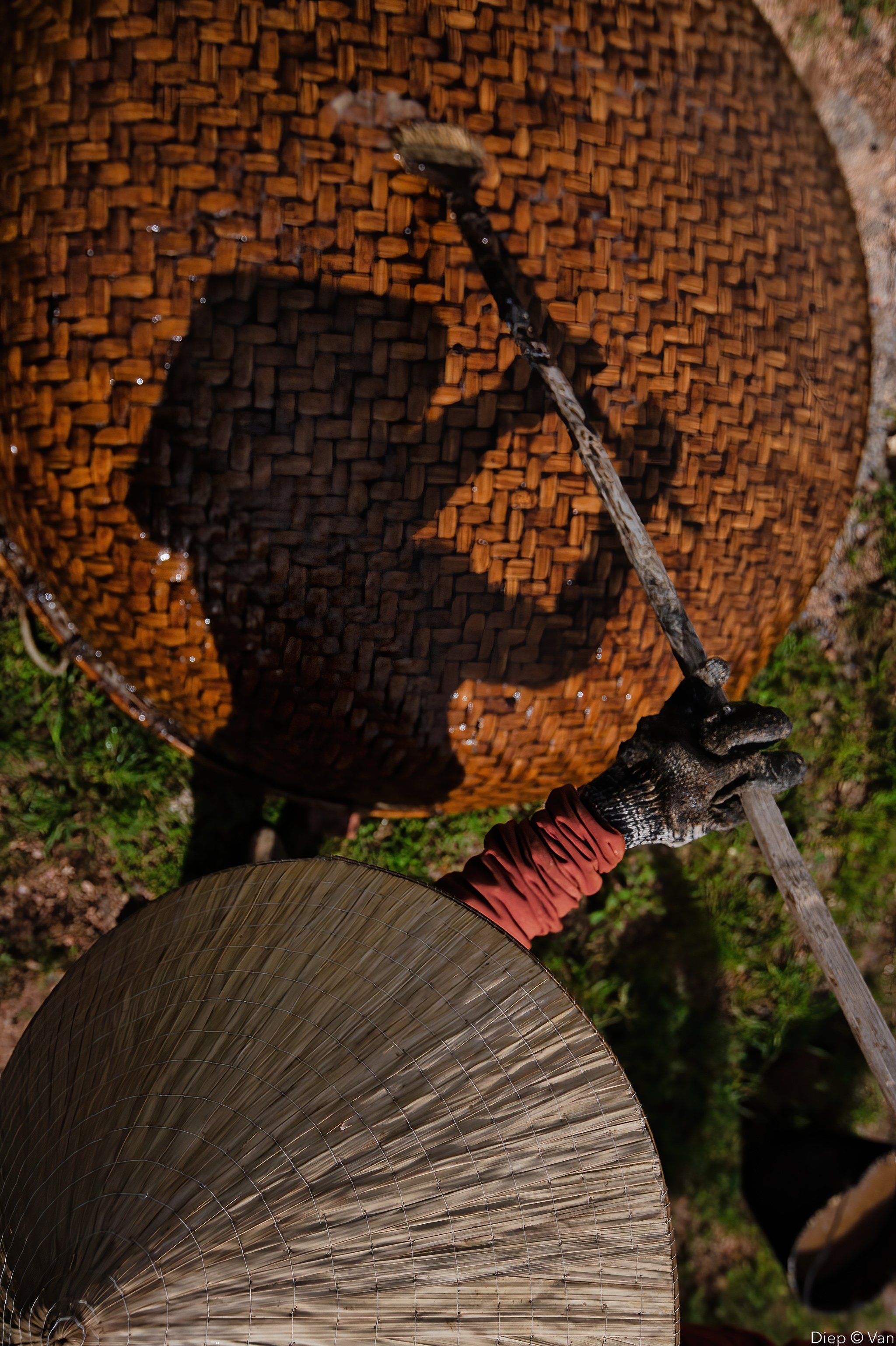 A person wearing a glove and red sleeve is holding a long stick and stirring a large, round, woven basket filled with water and small brown objects, possibly seeds or grains. The basket casts a shadow inside it, and an umbrella is visible at the bottom of the image, with a focused outdoor setting.