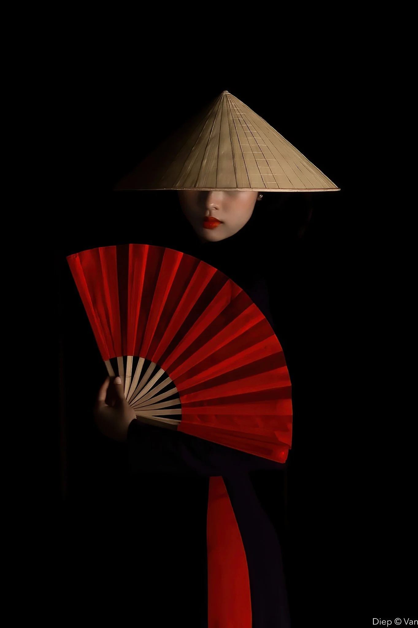 A woman wearing a conical Asian hat, holding a red folding fan, with her face partially visible against a dark background.