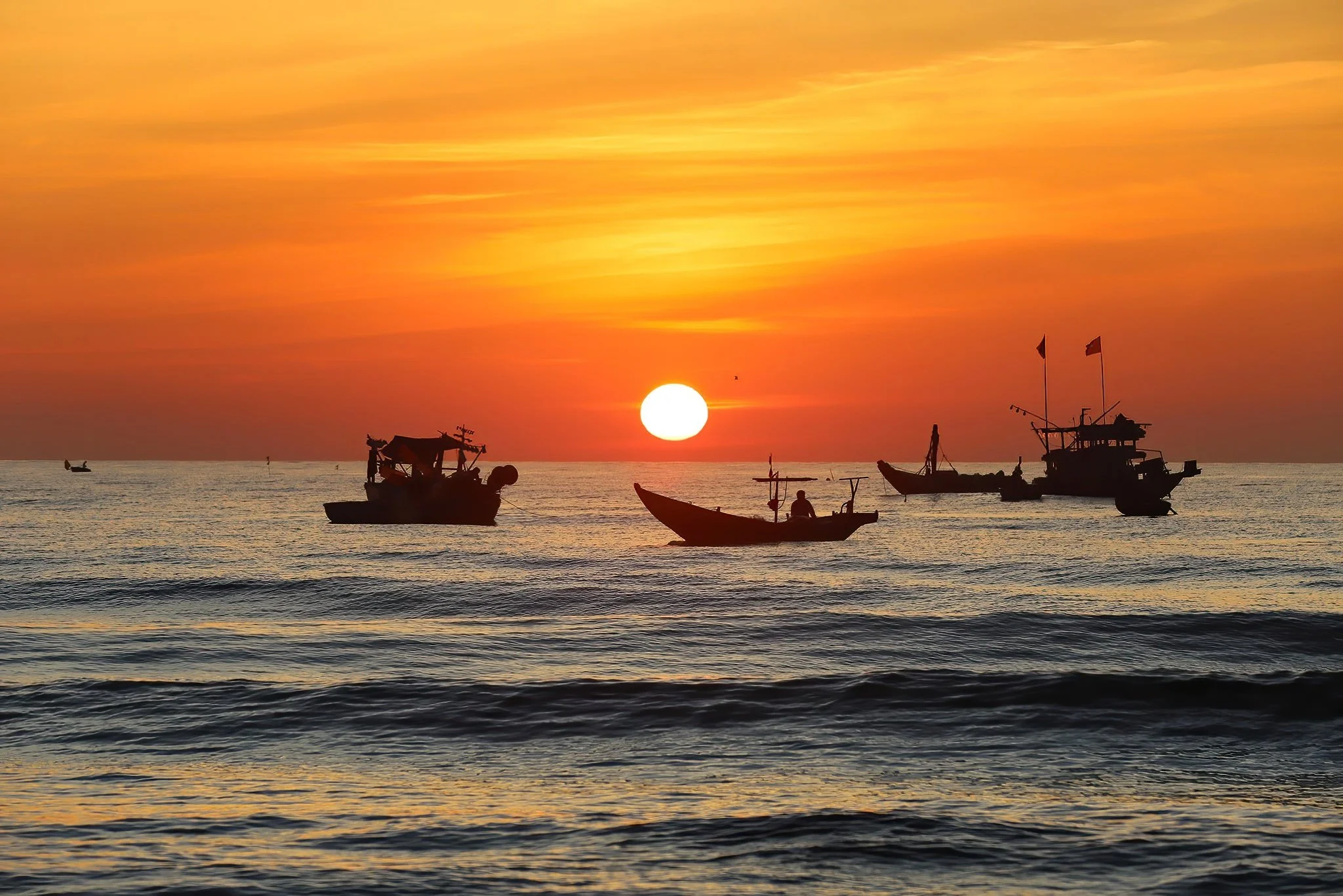 Silhouettes of several boats on the ocean during sunset with a colorful sky and a large, setting sun in the background.