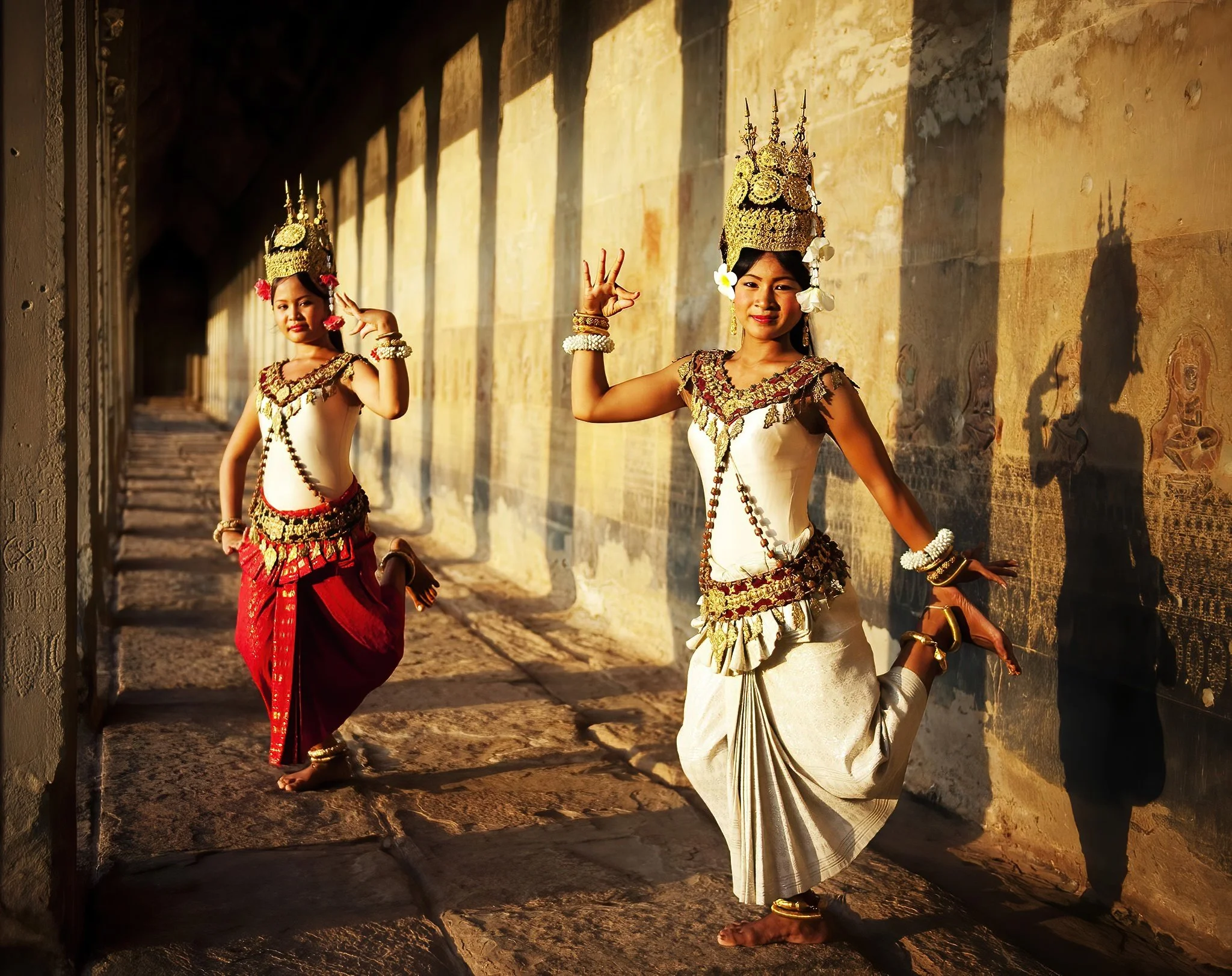 Two young girls dressed in traditional Cambodian dance costumes performing a dance in a historic temple corridor, with sunlight casting shadows on the wall.