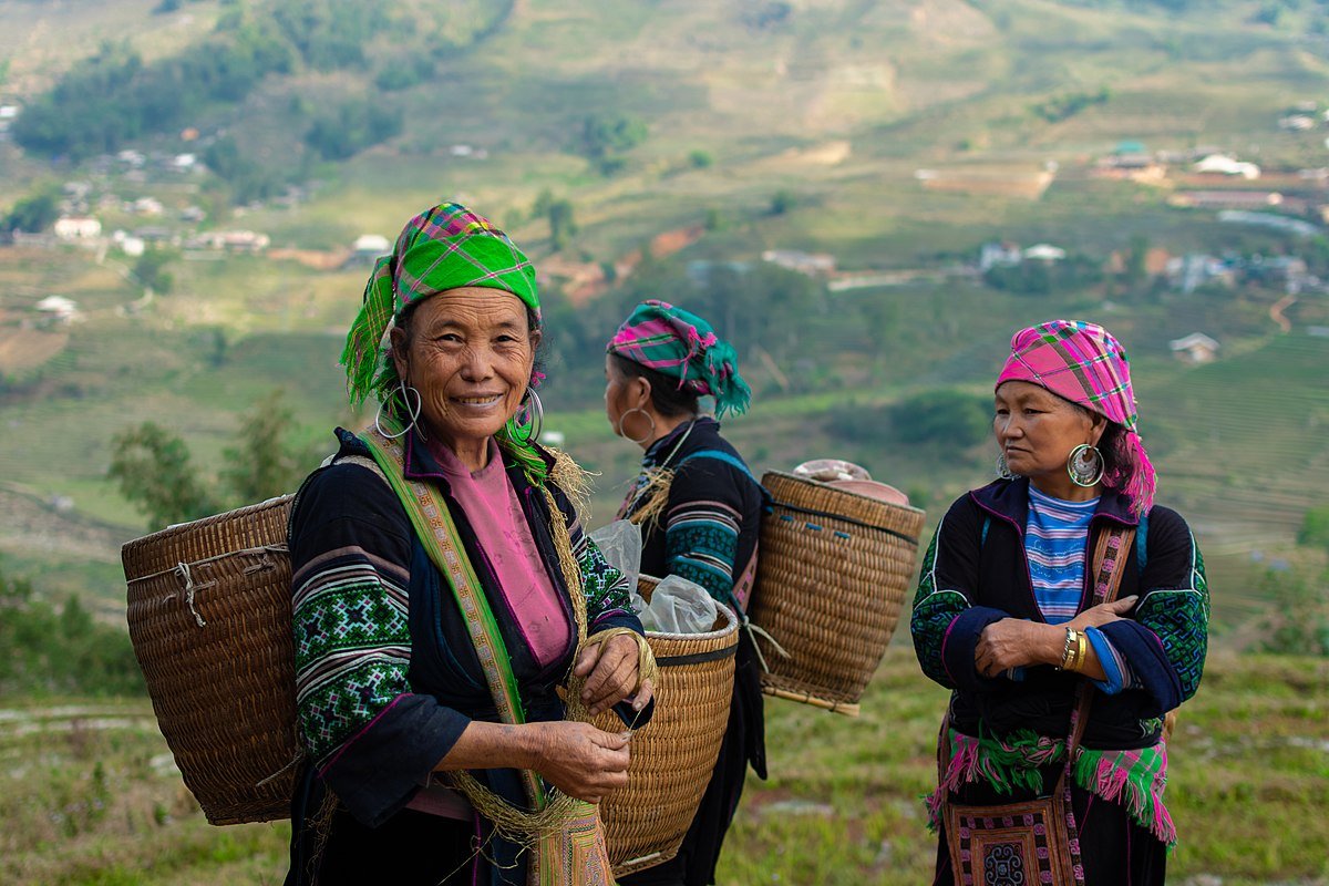 Three women dressed in traditional clothing, carrying woven baskets on their backs, standing outdoors in a hilly landscape with small houses in the background.