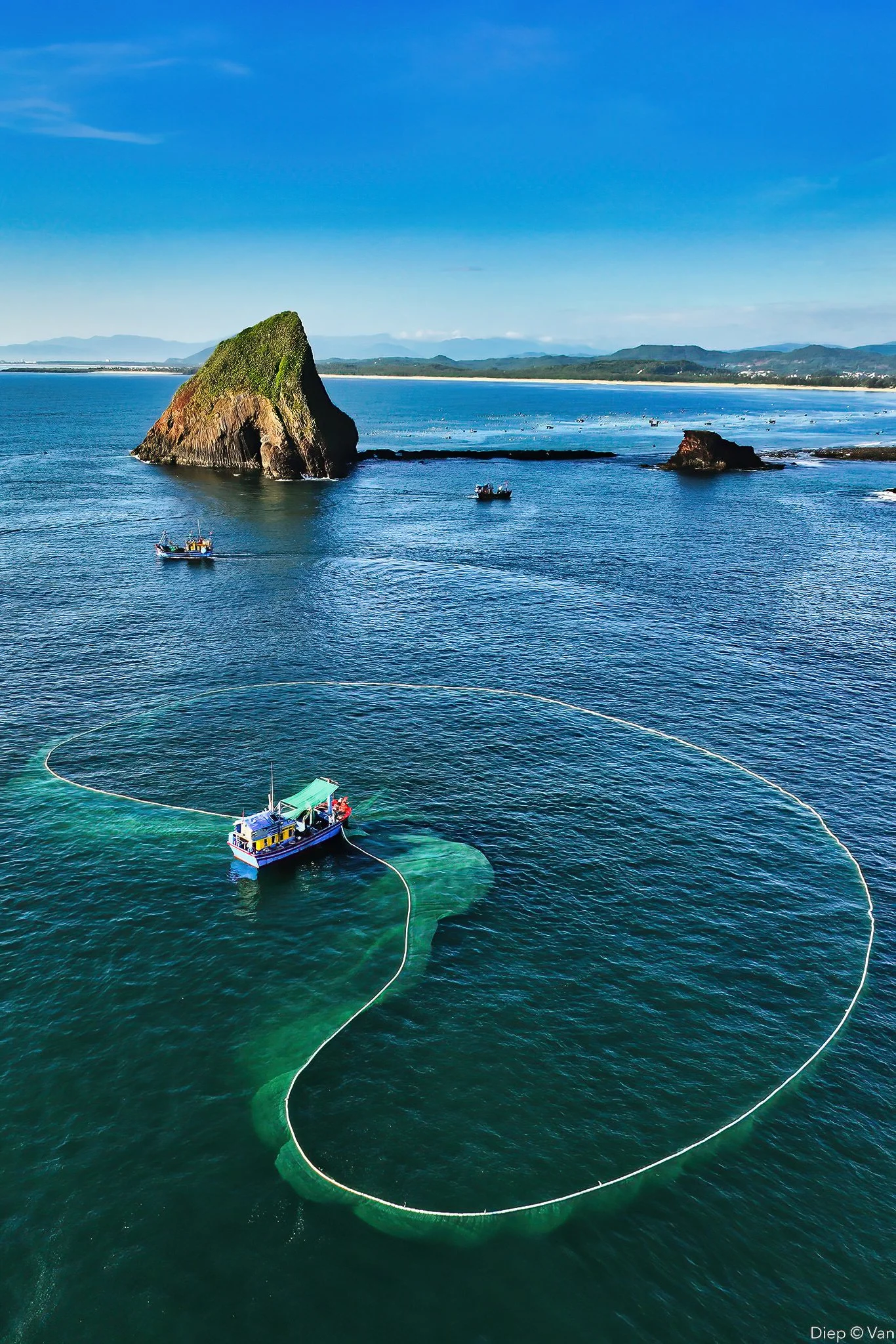 A boat in the ocean deploying a large fishing net in the shape of a heart, with a large rock formation and distant shoreline in the background.