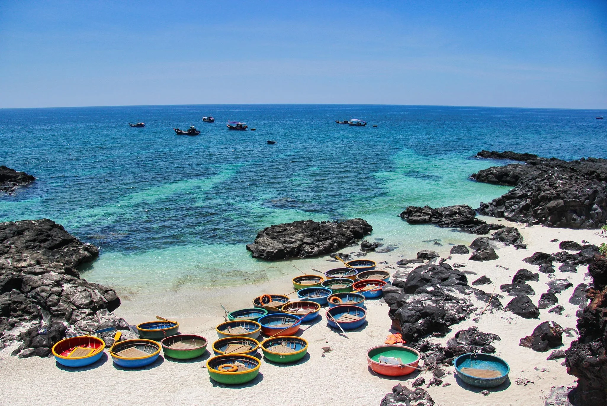 Colorful round boats resting on white sand beach with black rocks and a blue ocean in the background.