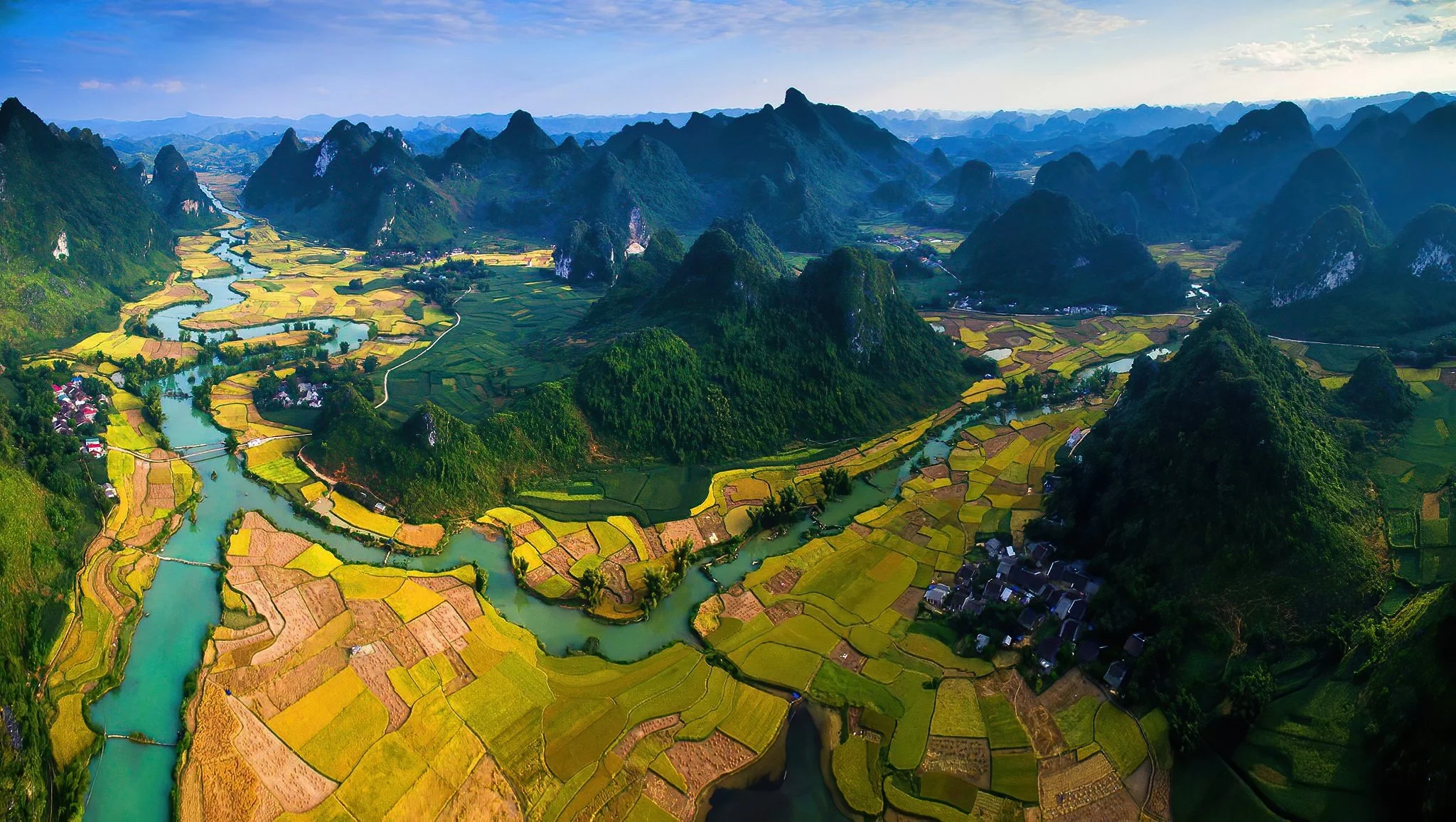 Aerial view of a lush green valley with winding river, surrounding karst mountains, patchwork of yellow and green farmland, and small villages.