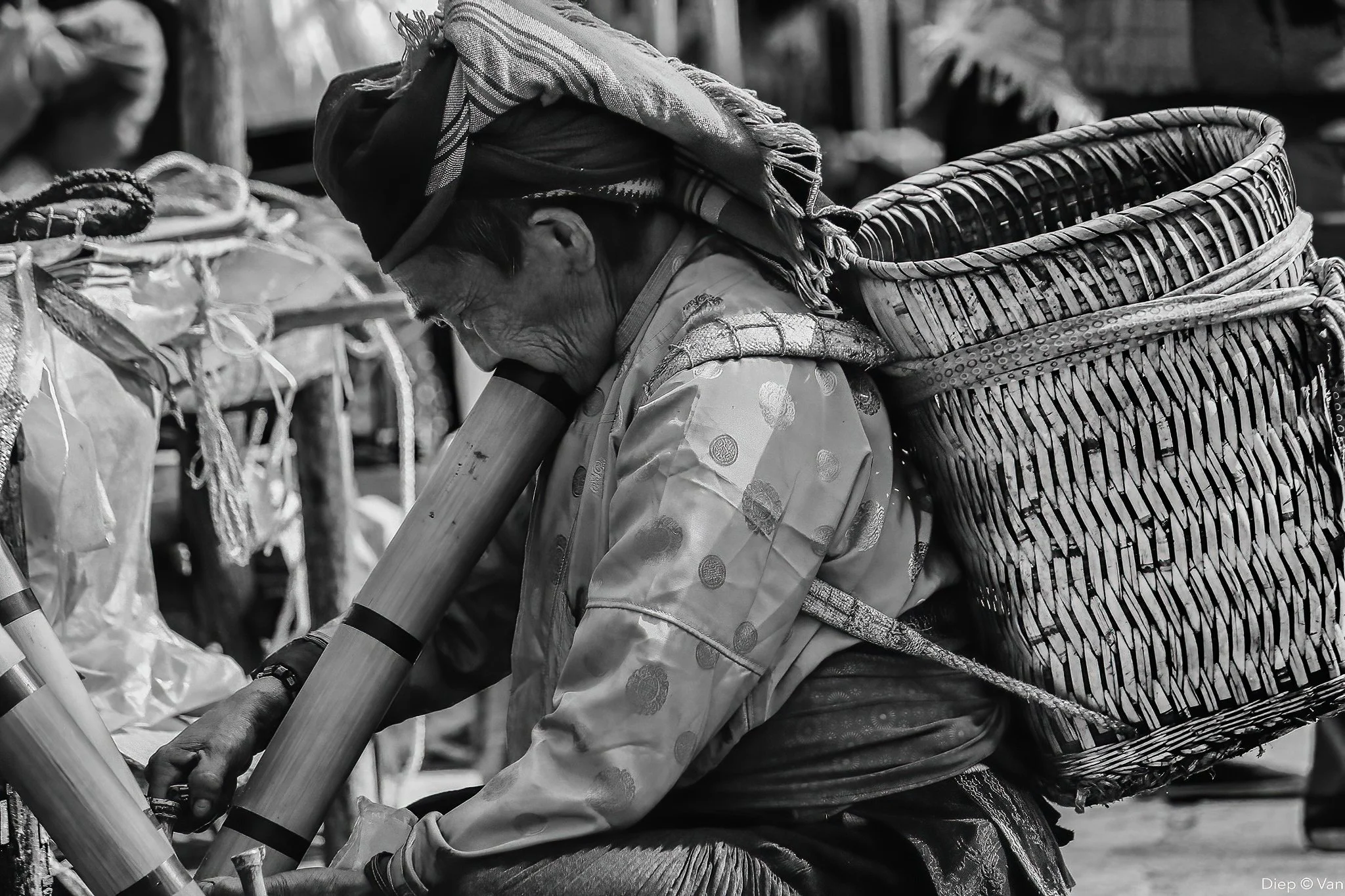 An elderly woman wearing traditional clothing and headscarf, carrying large woven baskets on her back, working at an outdoor market in black and white.