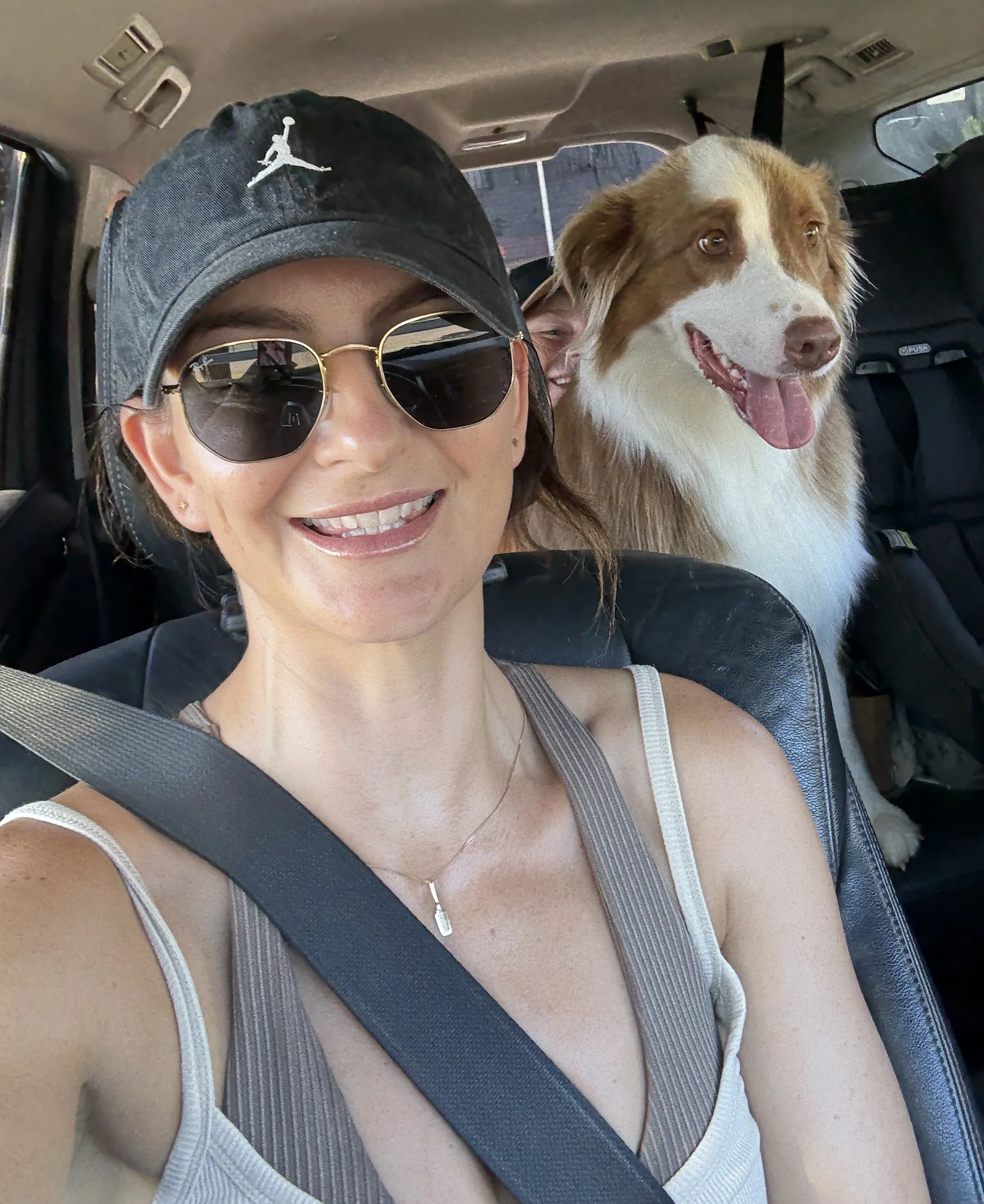 A woman wearing sunglasses and a baseball cap smiling while sitting in a car with a smiling dog behind her.