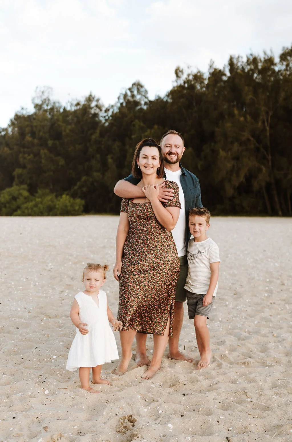 A happy family of four standing on a sandy beach with trees in the background, smiling at the camera. The father has his arm around the mother, who is wearing a floral dress. The son stands next to the mother, and the daughter is standing in front, wearing a white dress.