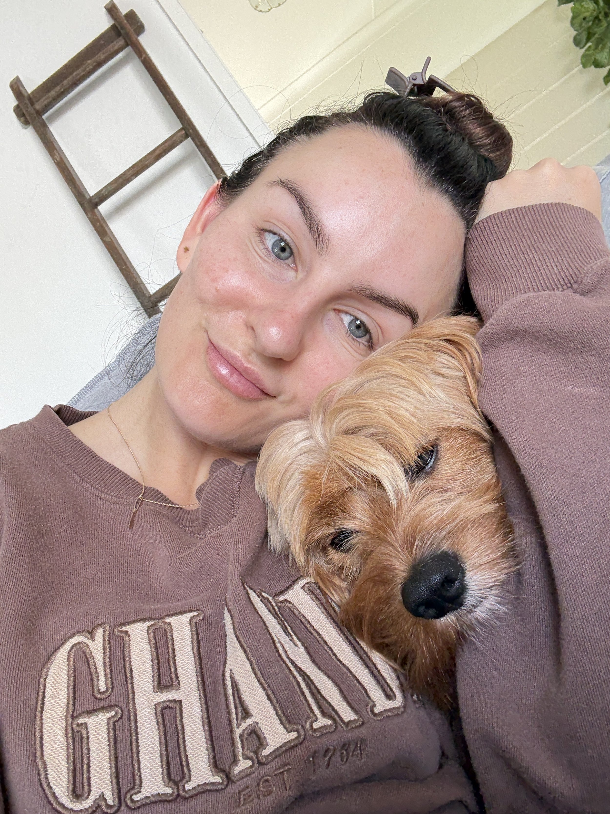 A young woman with dark hair pulled back, lying down with her head resting on her arm, smiling at the camera. She is wearing a brown sweatshirt with the word 'CHAMP' on it. Next to her, a small, light brown dog with shaggy fur is resting its head on her shoulder, partially covering her. The background shows part of a wall, a wooden ladder, and a potted plant.