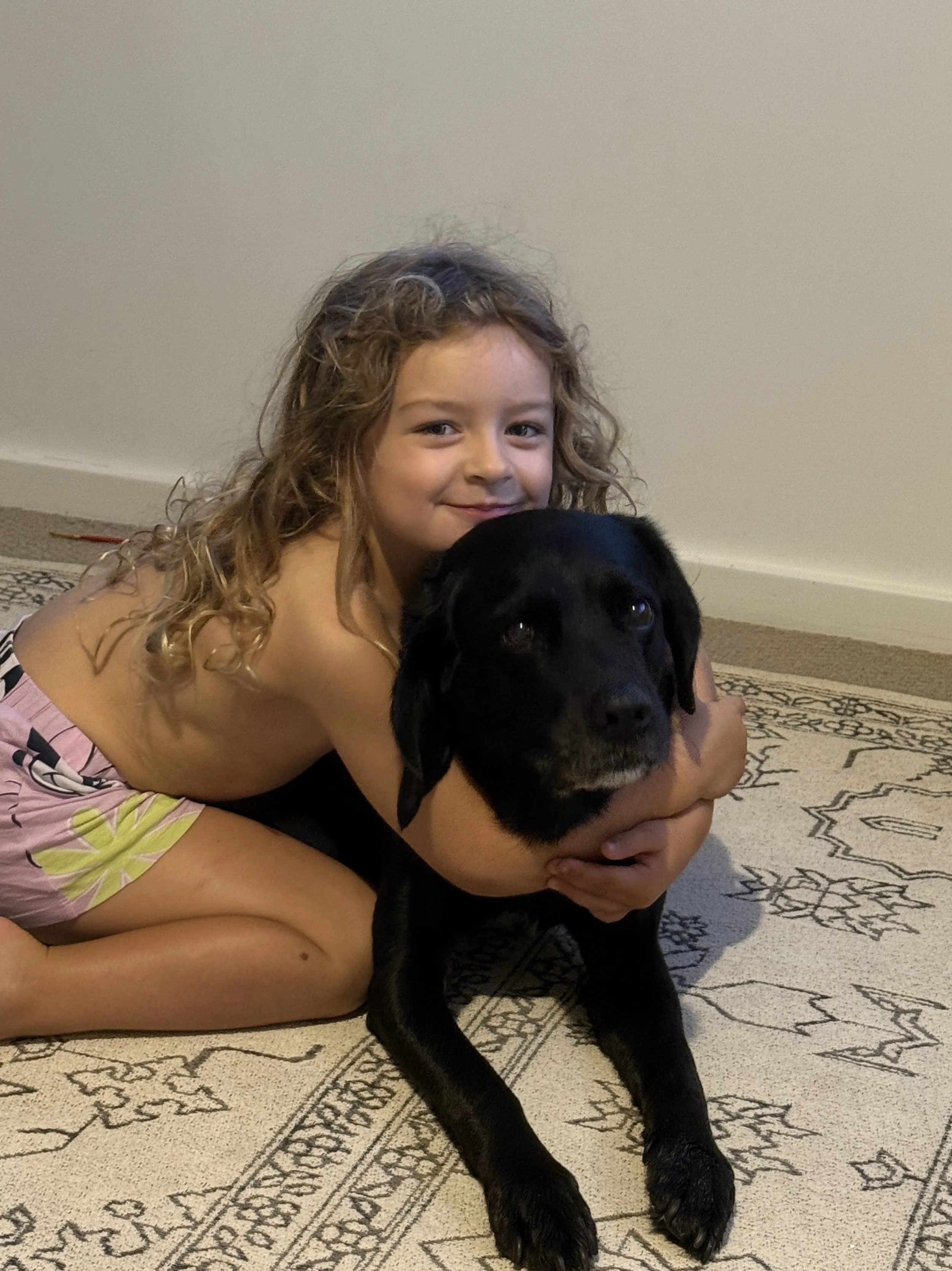 A young girl with curly brown hair hugging a black Labrador Retriever puppy on a patterned beige rug inside a home.