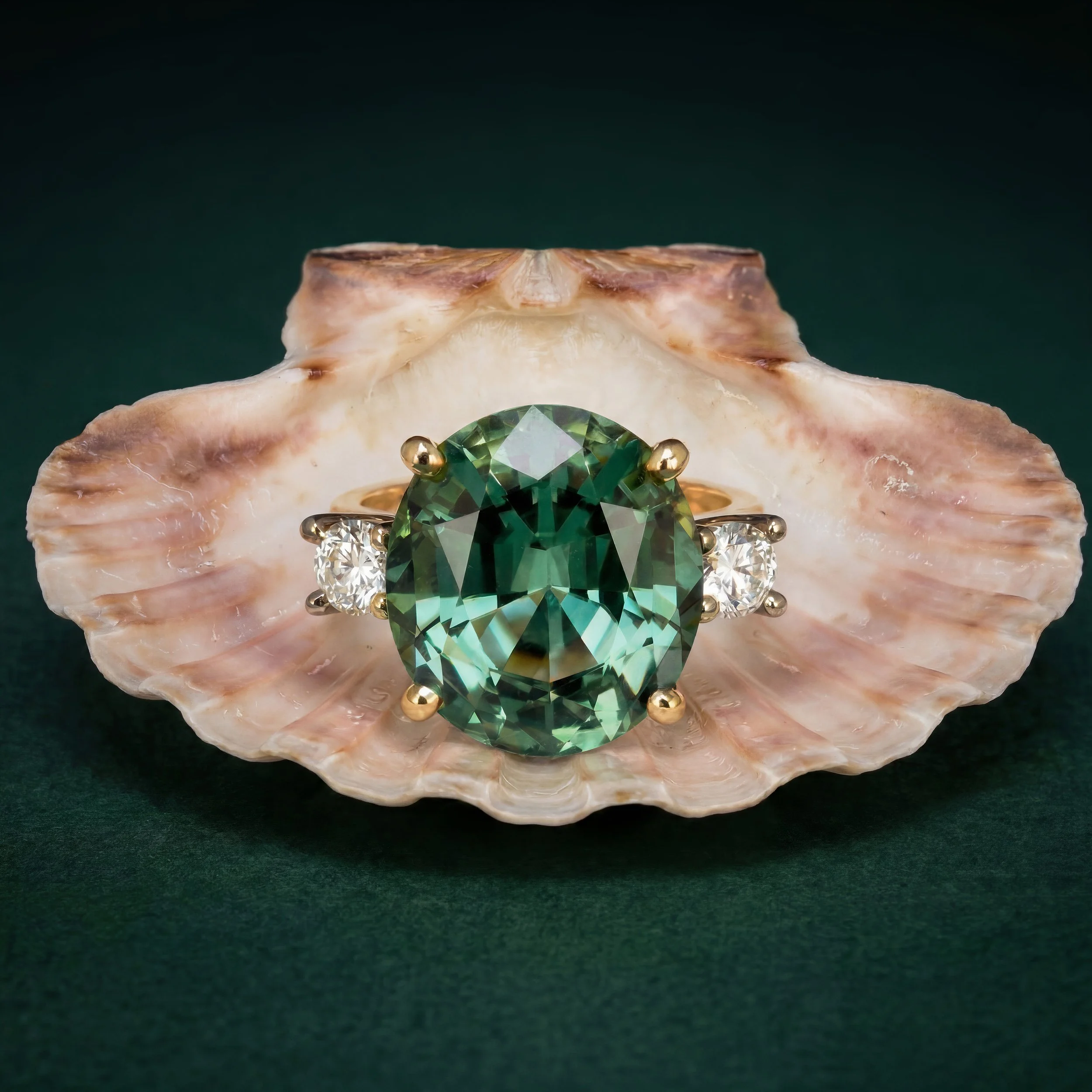 Close-up of a green gemstone ring set in gold, placed in an oyster shell with rough pink exterior, against a dark green background.