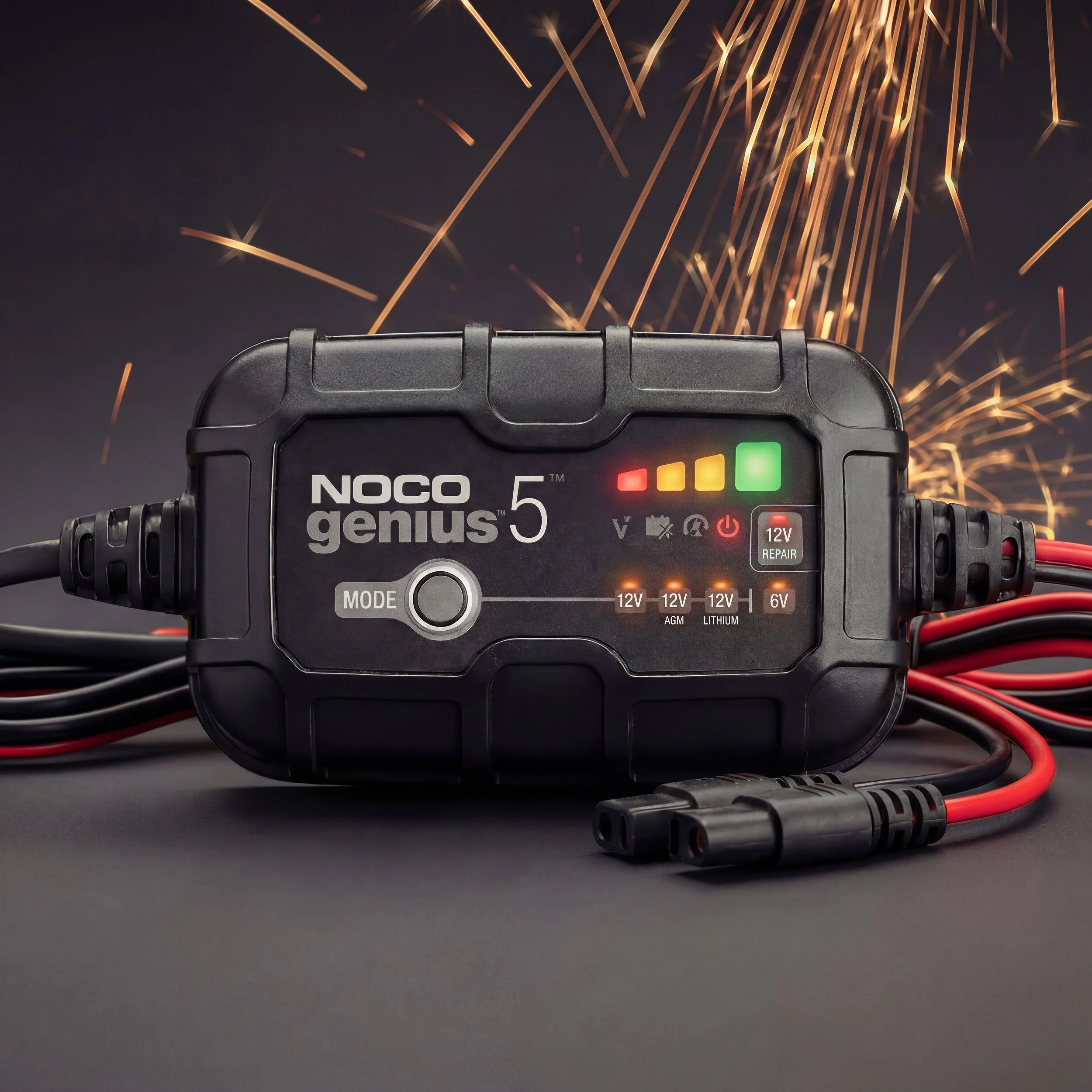 Clost-up of a NOCO Genius 5 battery charger with red and black cables attached, sparks flying in the background.