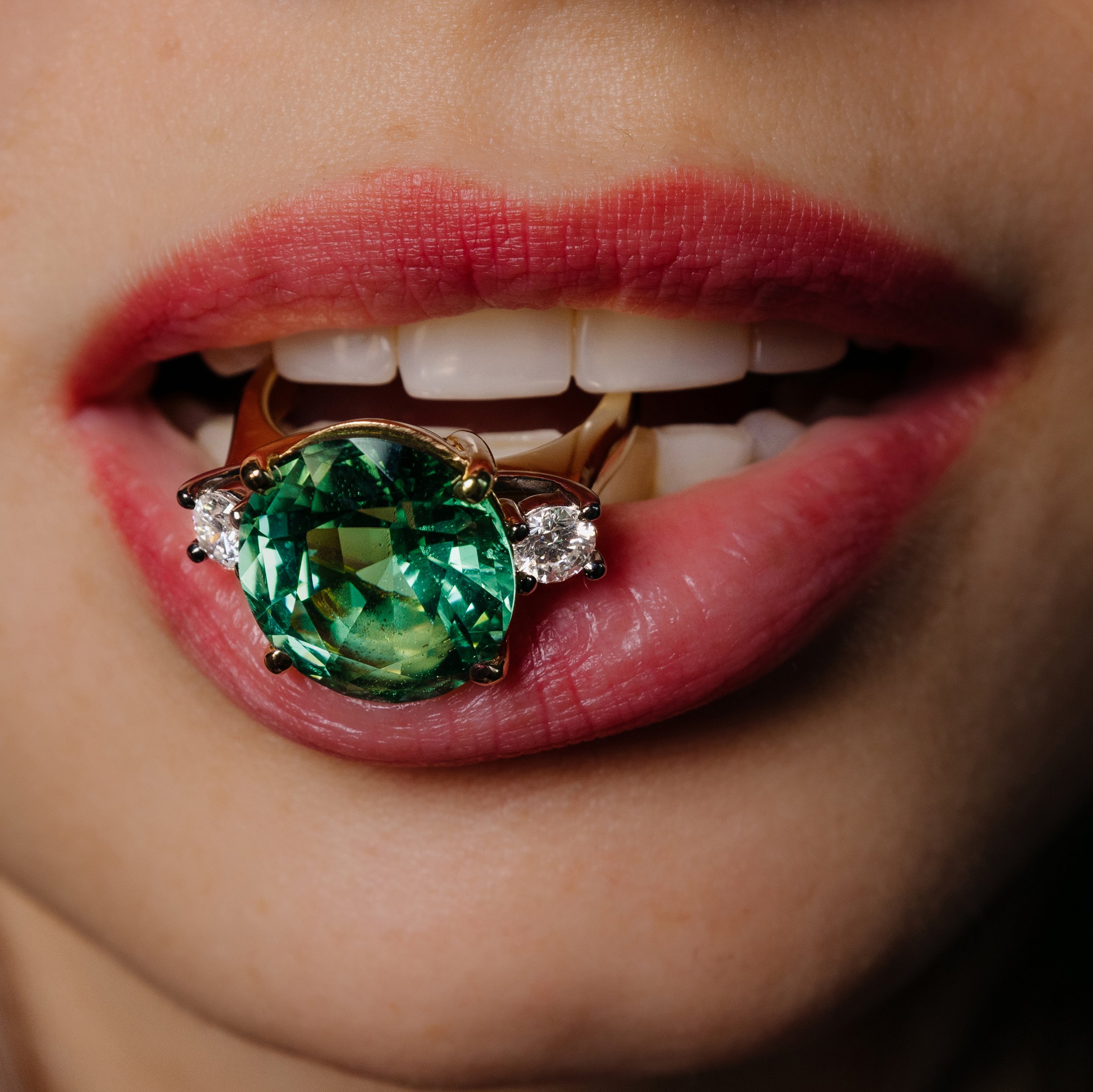 Close-up of a person's lips with red lipstick, jewelry with a green gemstone, teeth, and part of their face.