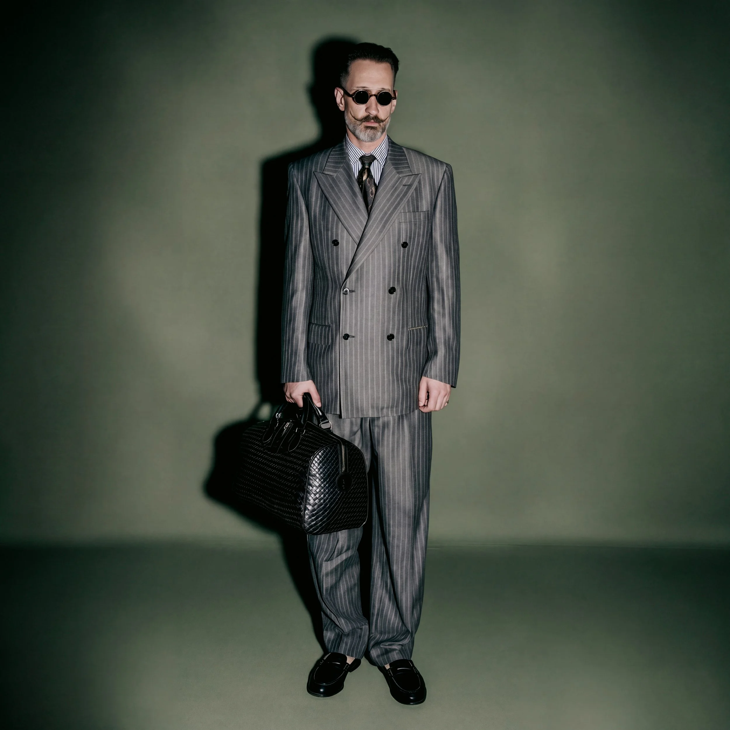 A man in a gray pinstripe double-breasted suit, wearing dark round sunglasses, a striped shirt, and a patterned tie, stands against a green background. He has a gray beard and holds a black quilted handbag in his left hand, with his right hand hangin
