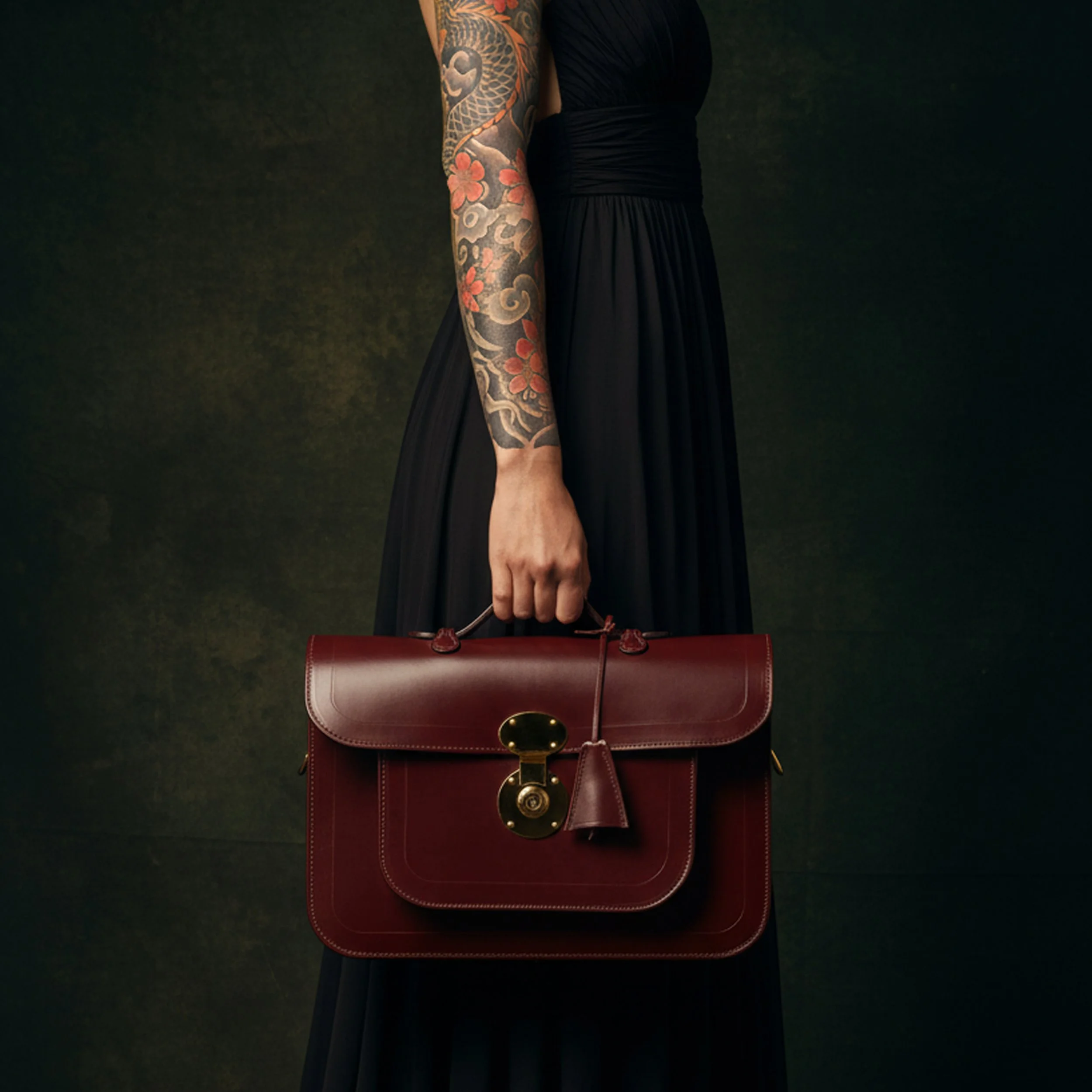 A woman wearing a black dress with a colorful tattoo on her arm, holding a red handbag with gold hardware, standing against a dark background.