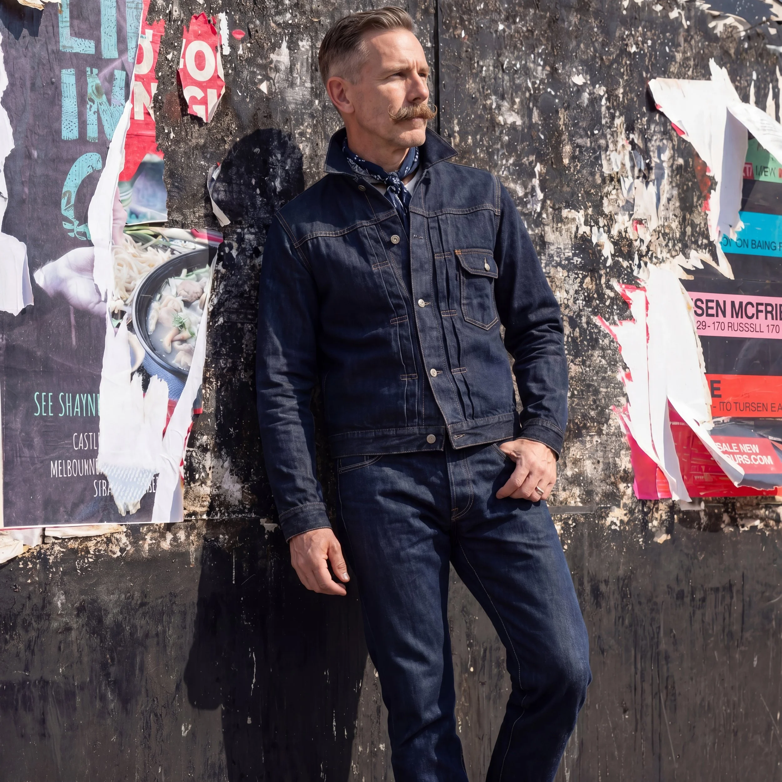 A man with a mustache wearing a denim jacket and a blue scarf stands outdoors against a black textured wall with torn posters and graffiti.