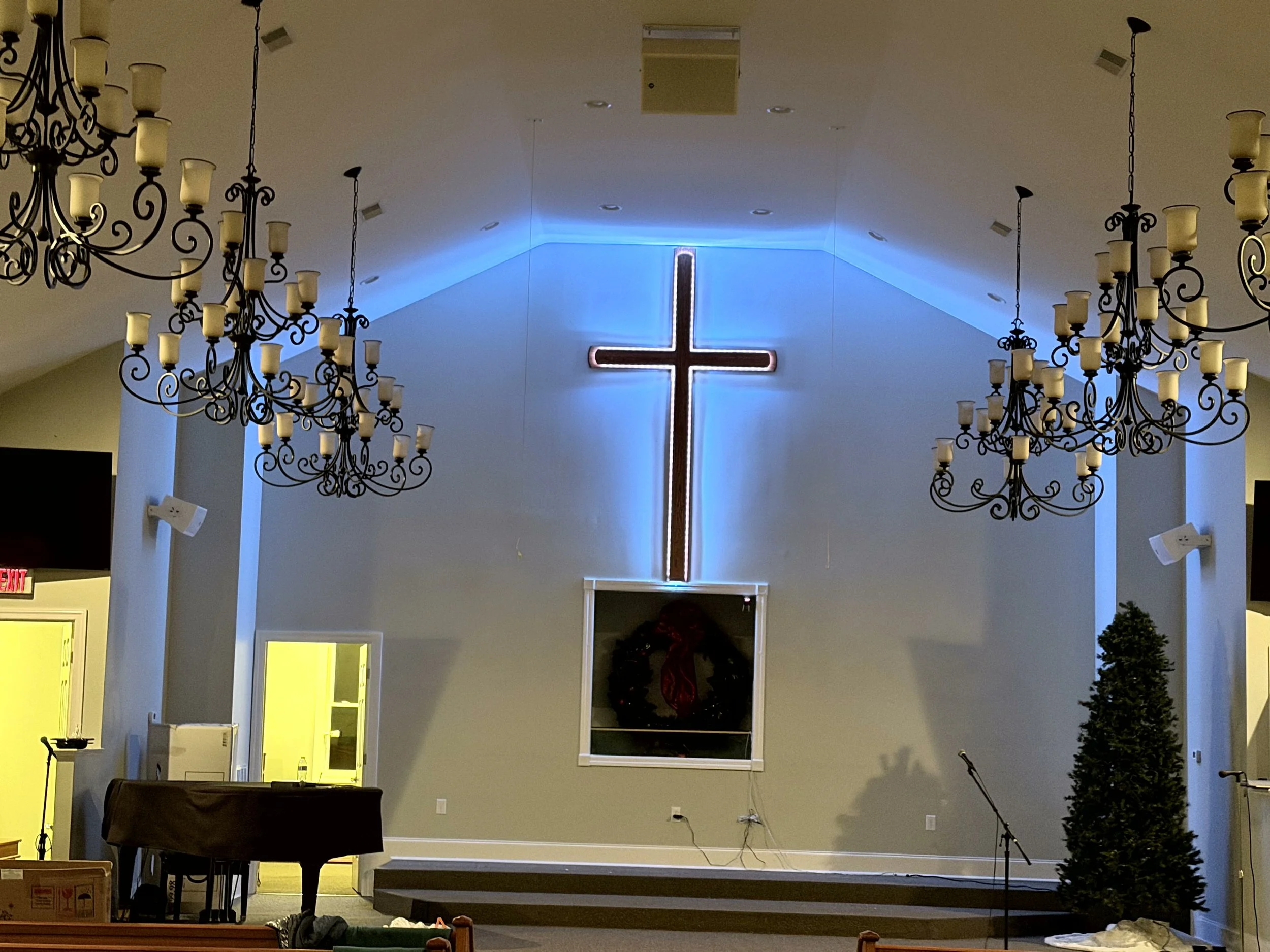 Interior of a church with a large illuminated cross on the wall behind the stage, two chandeliers hanging from the ceiling, a small Christmas tree on the right, and a wreath on the wall below the cross.
