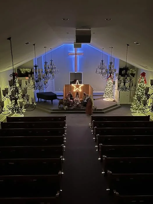 Interior of a church decorated for Christmas with Christmas trees and a cross illuminated in neon blue on the wall behind the altar, with people gathered.