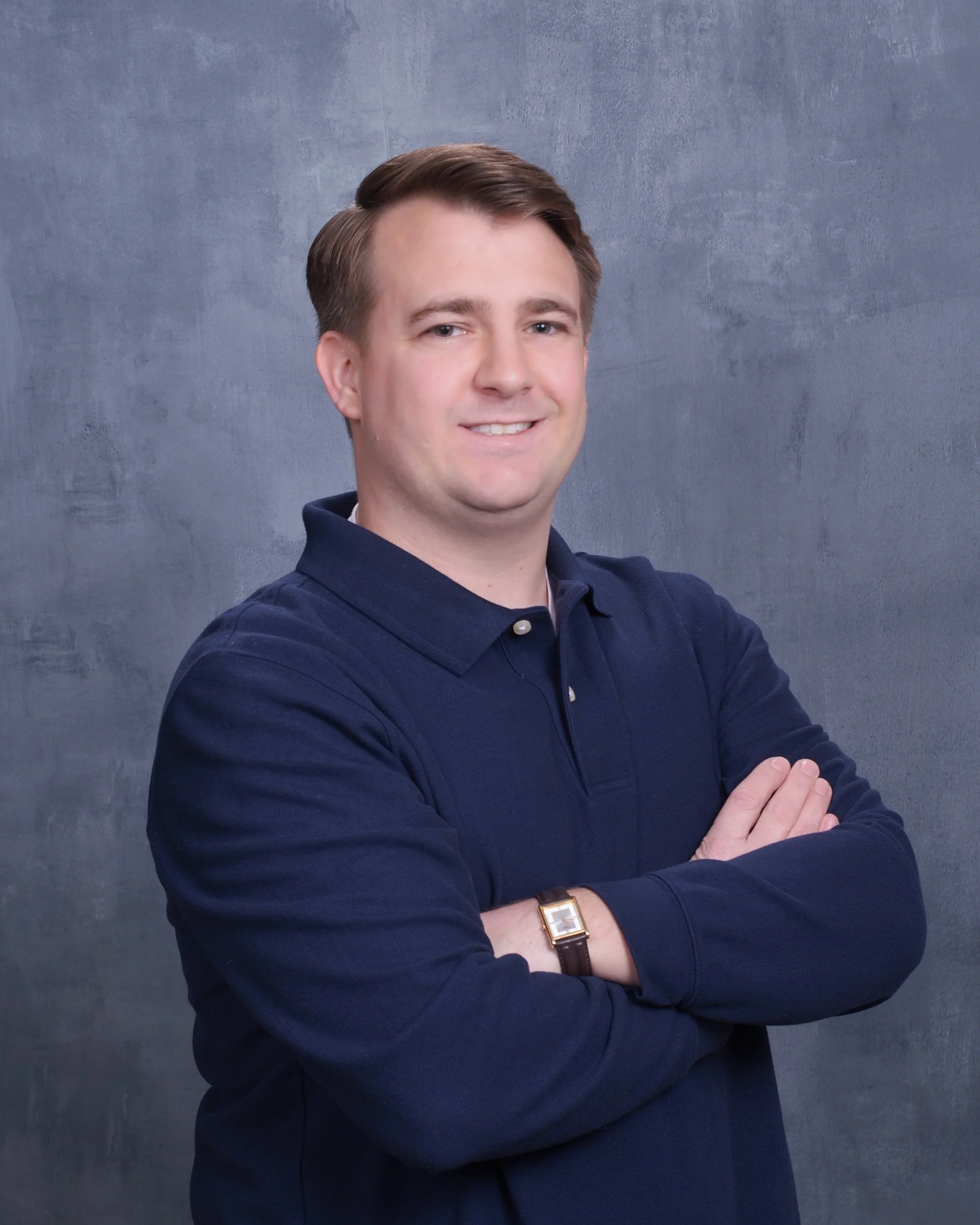 A man with brown hair wearing a navy blue collared shirt, crossing his arms, standing in front of a textured gray background.