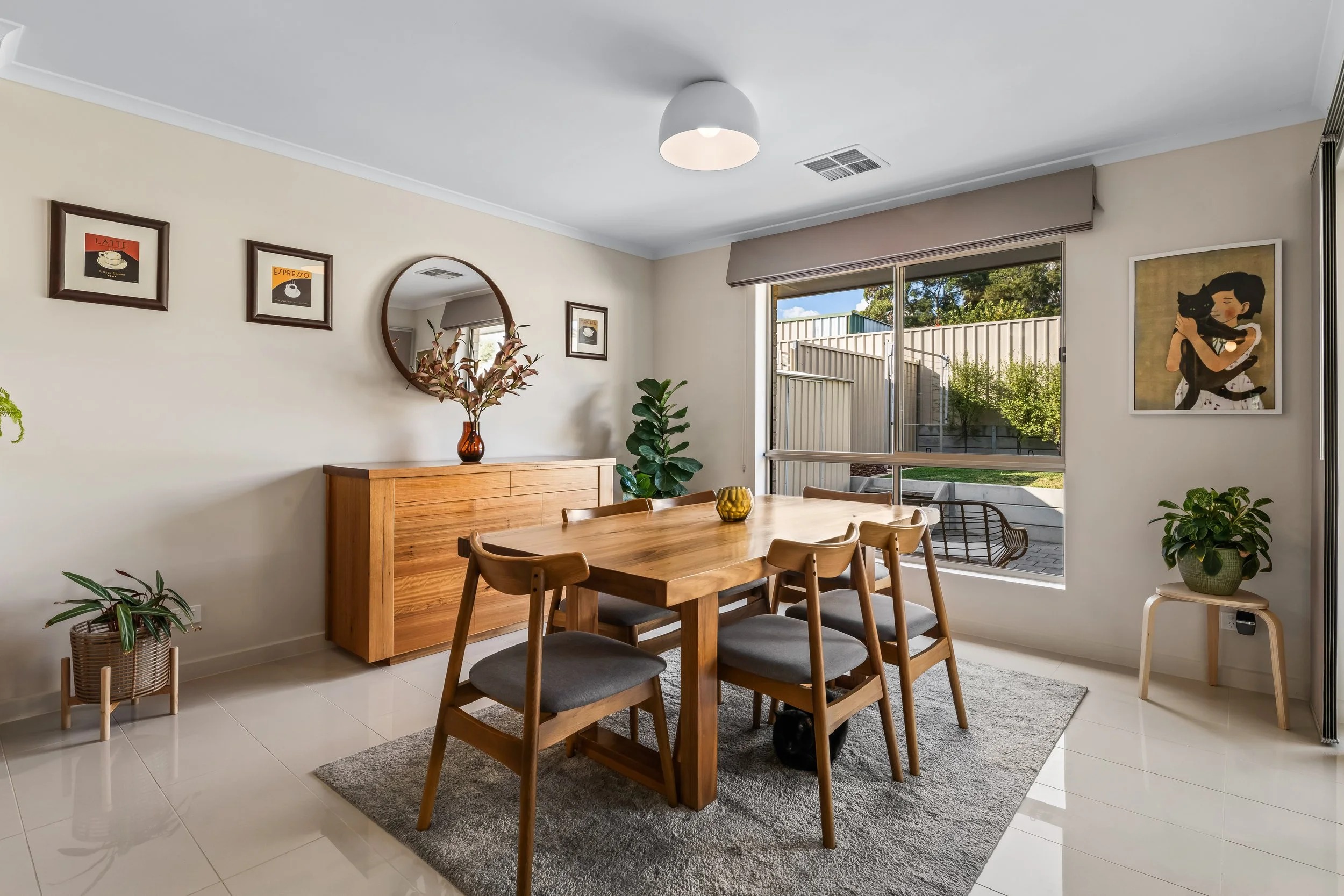 Interior of a dining room with a wooden table and six chairs, a wooden sideboard, potted plants, framed artwork on the wall, a large window with a garden view, and a ceiling light.