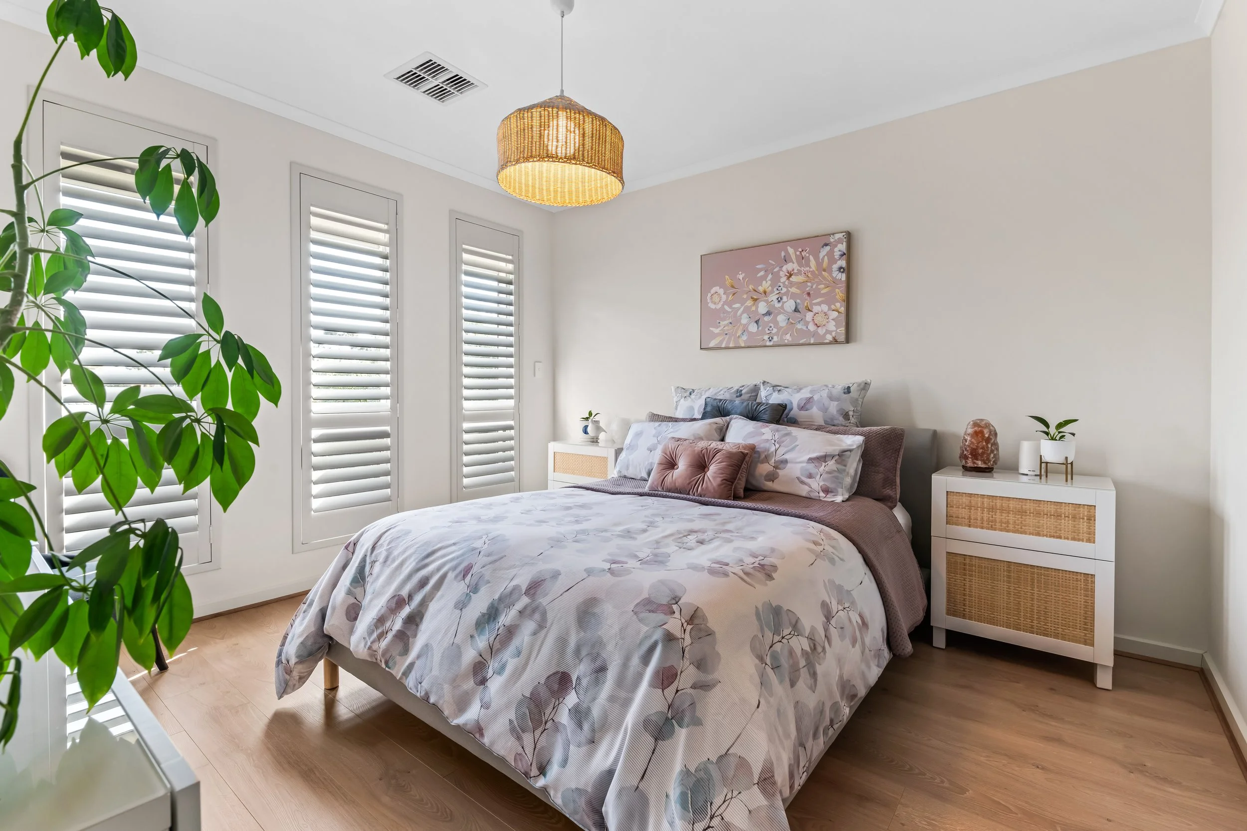 A bedroom with a bed covered in floral bedding, a white bedside table with a salt lamp, indoor plants, and three windows with white blinds, decor includes a pink floral painting and a rattan lampshade.