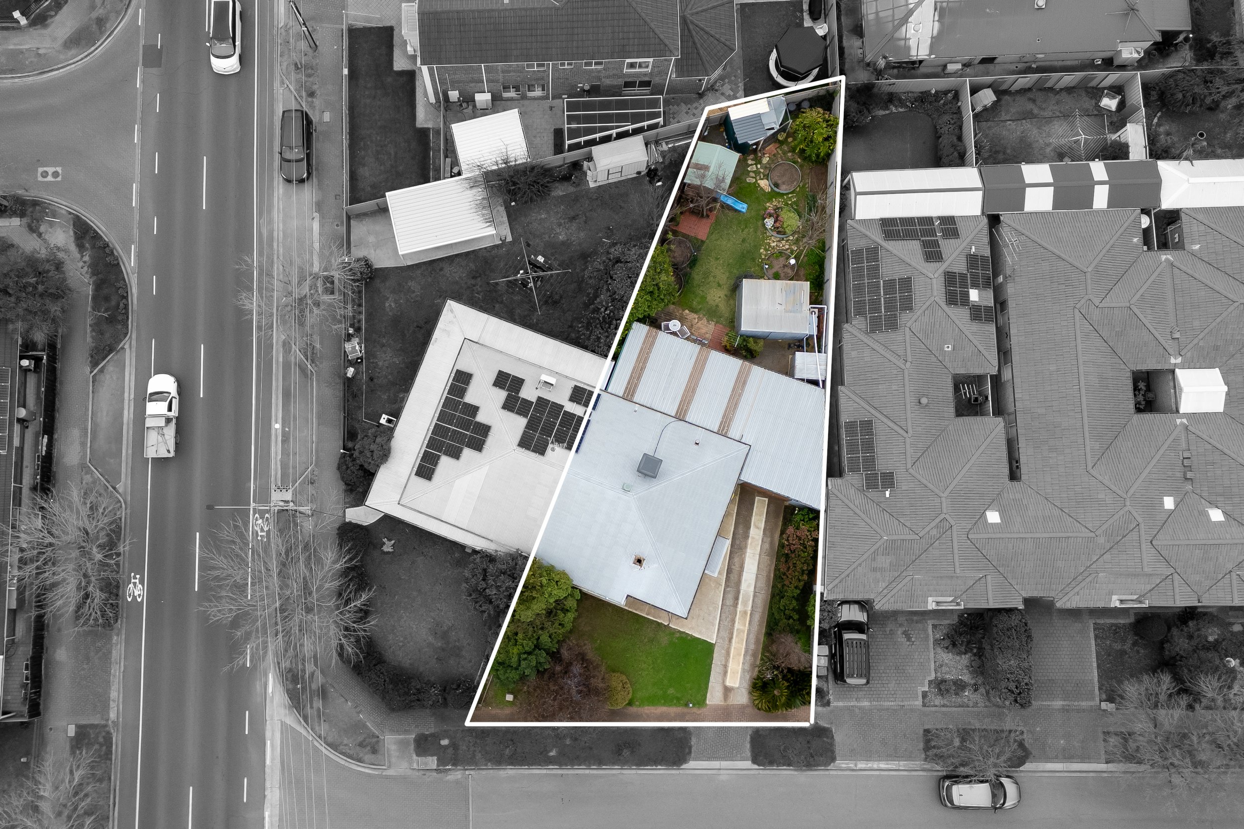 Aerial view of a residential area showing a backyard with a small garden, trees, and a play area. The yard is outlined with a white border. Surrounding the yard are neighboring houses with tiled roofs, some of which have solar panels. A street with c