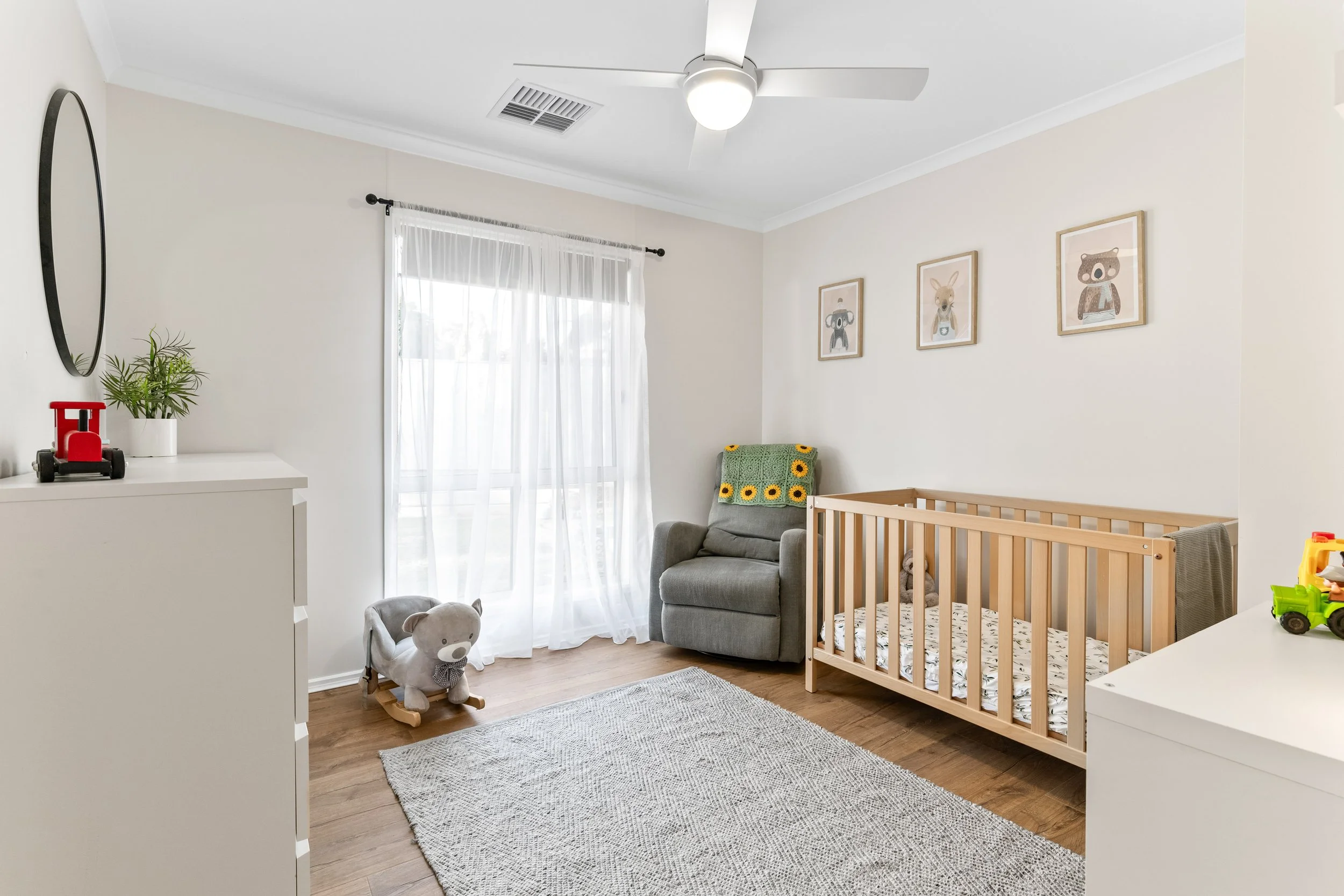 A baby nursery with a crib, a plush teddy bear, a grey armchair with a sunflower blanket, wall art of animals, a white dresser with a plant and a toy truck on top, sheer curtains on a window, and a ceiling fan with a light.