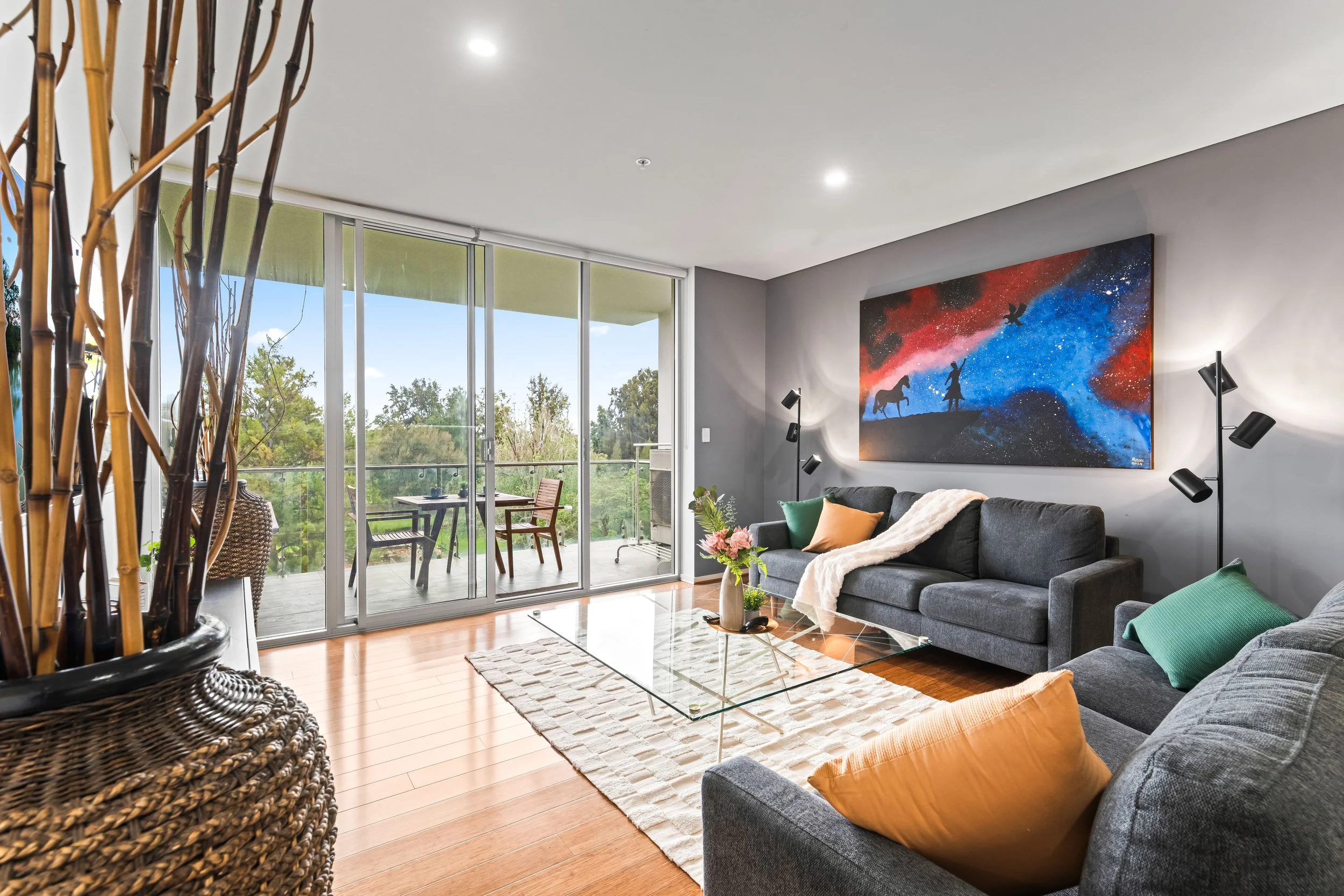 Living room with large glass sliding doors opening to a balcony and a view of trees outside. The room contains a dark gray sofa with colorful pillows, a glass coffee table, a white rug, and a wall mounted artwork of a space scene with astronauts, pla