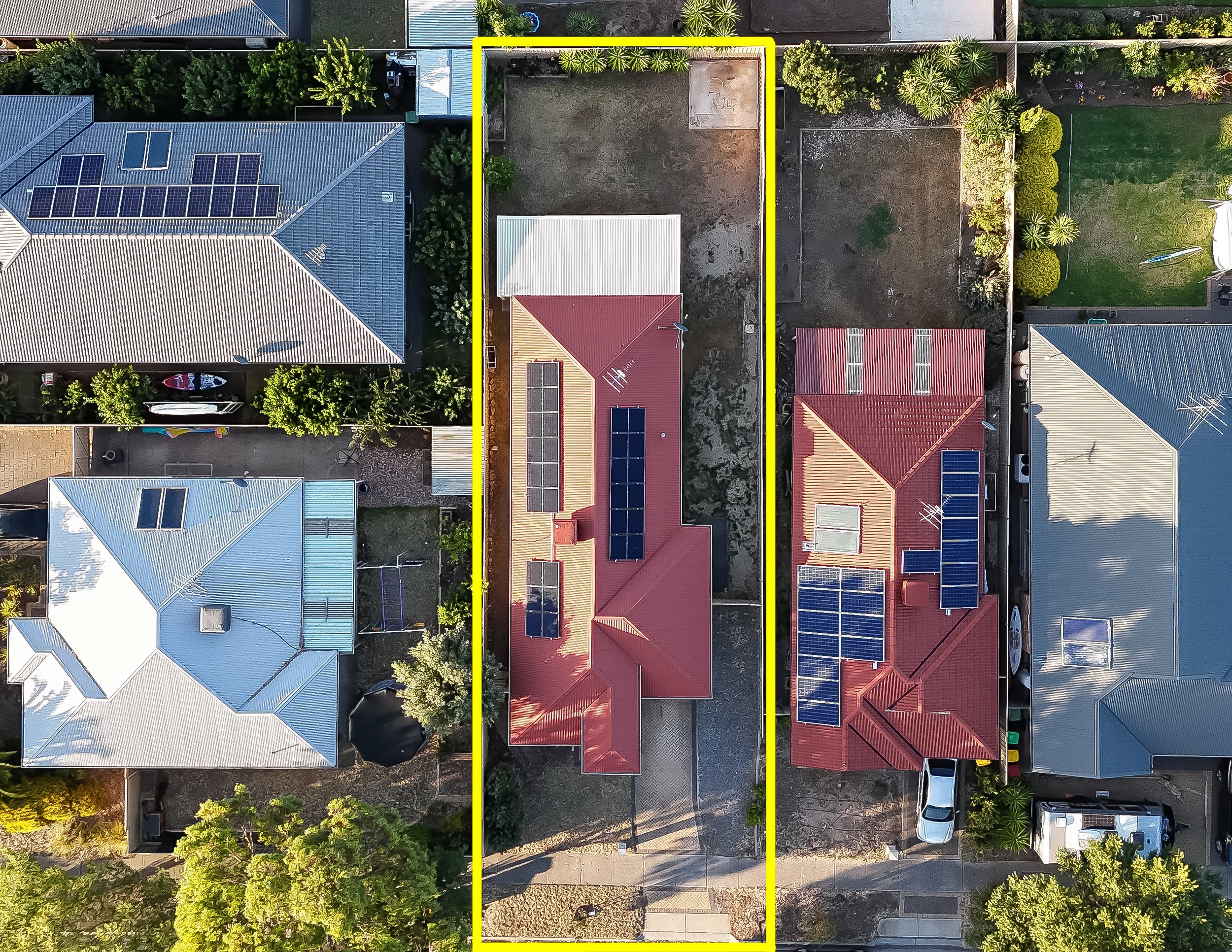 An aerial view of several houses with solar panels on their roofs on a suburban street. One house with a red roof, another with a blue roof, and another with a gray roof can be seen. The backyard of the house outlined in yellow is mostly empty soil w