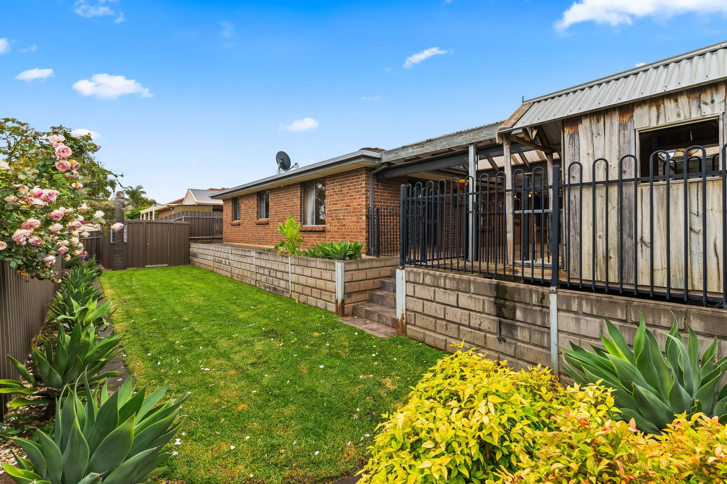 Backyard garden with green grass, flowering bushes, and a brick and wooden house with a fenced porch under a blue sky with clouds.