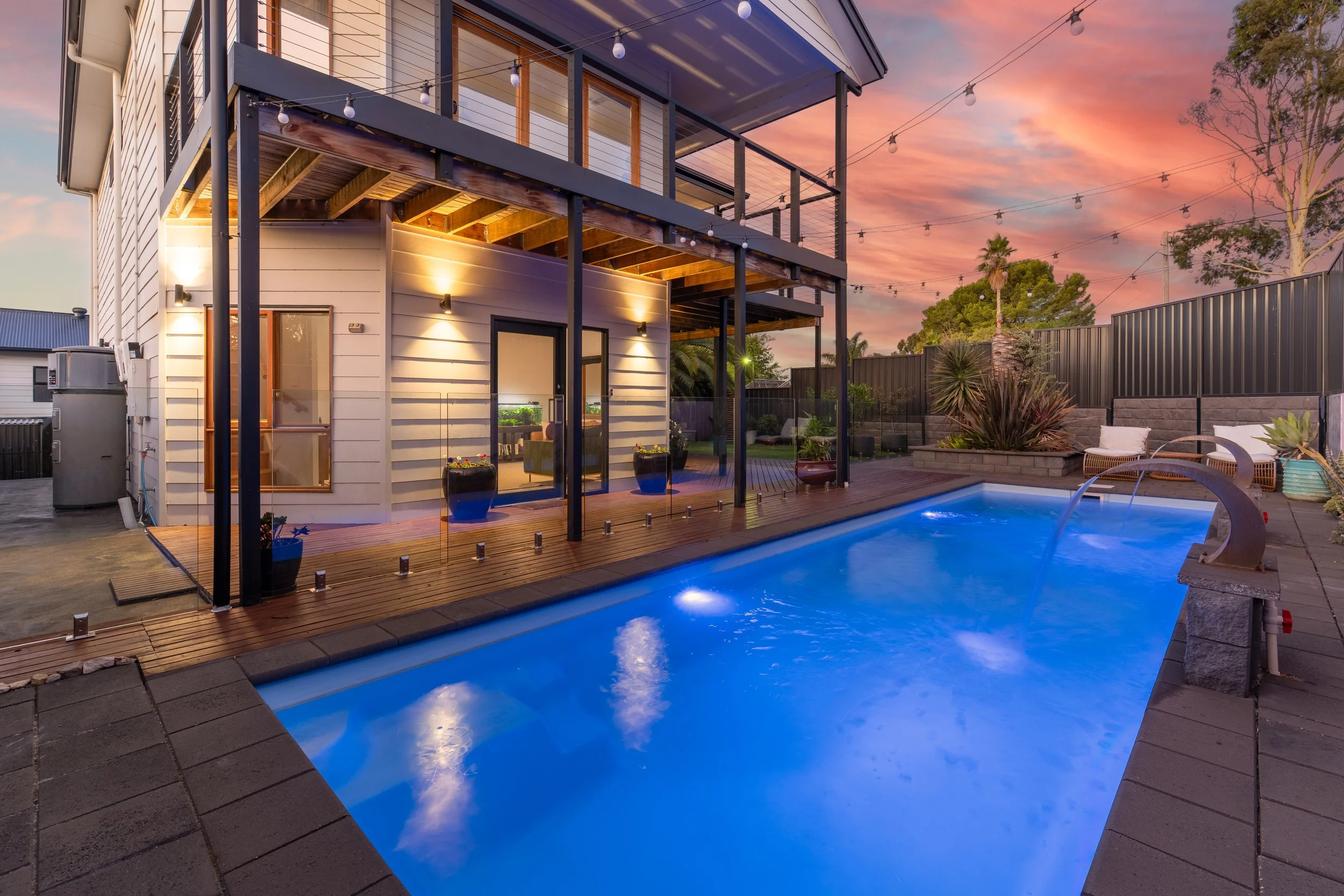 A backyard swimming pool at sunset with outdoor furniture, potted plants, and string lights hanging above, adjacent to a two-story house with an upper balcony.