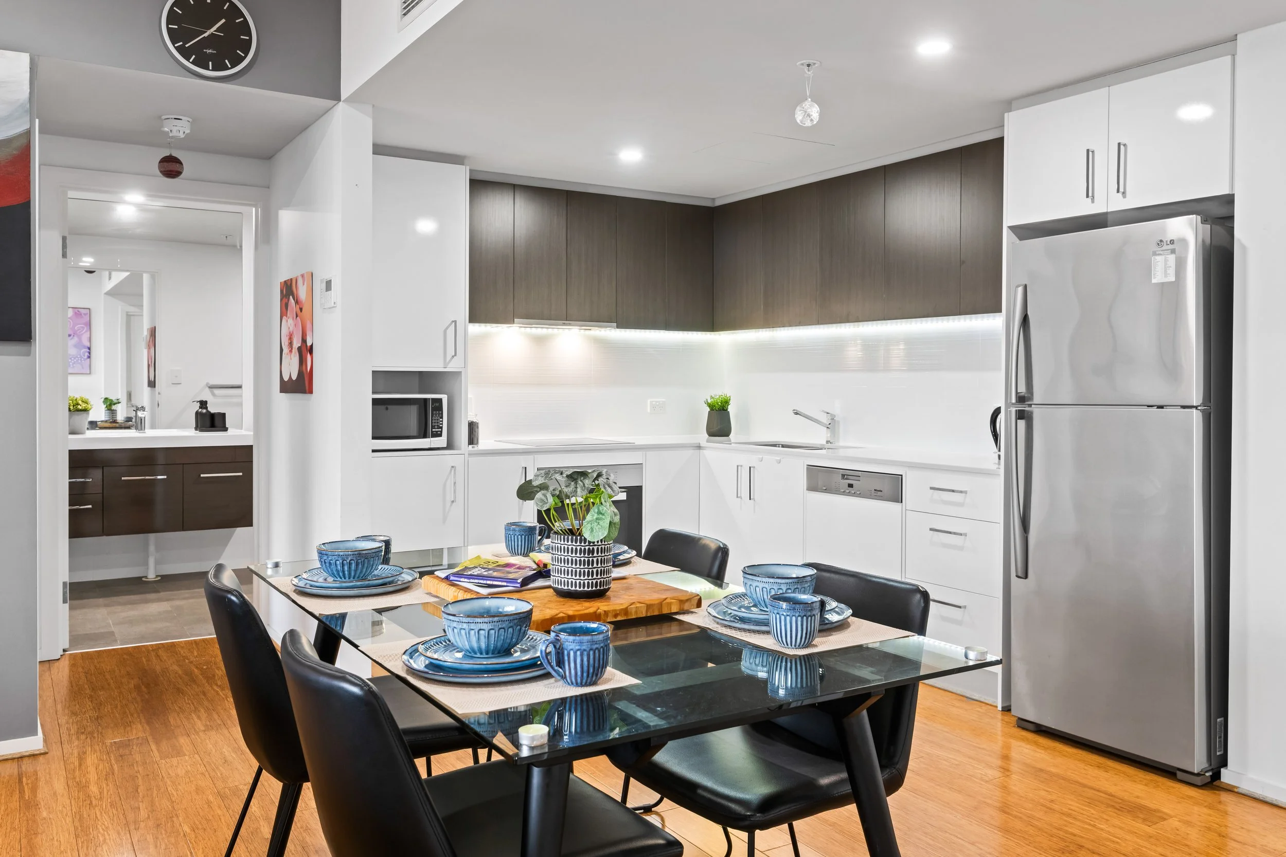 Modern kitchen with white cabinets, dark wood upper cabinets, stainless steel refrigerator, microwave, and a decorated dining table with blue dishes and a plant in the center.