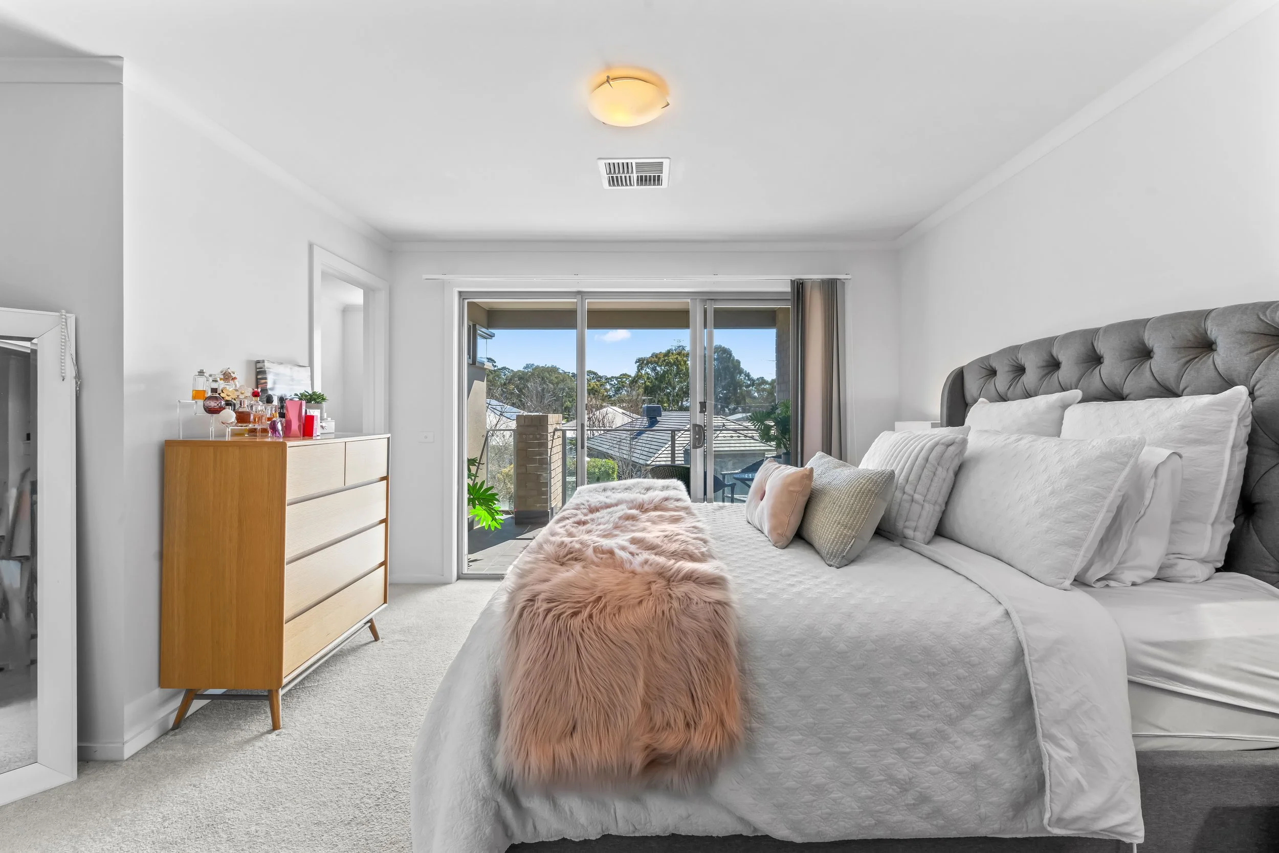 A bedroom with a large bed covered in white bedding and pillows, two pink fur accent throws at the foot of the bed, a gray tufted headboard, sliding glass door leading to a balcony with trees outside, a wooden dresser with perfume bottles and a potte