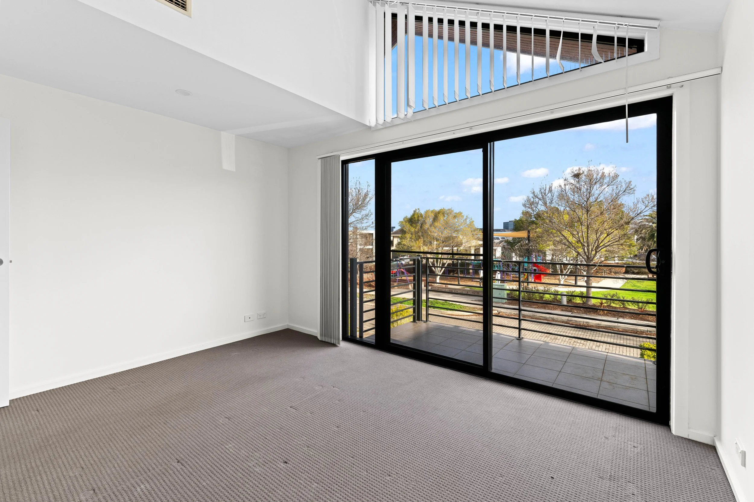 Empty living room with large sliding glass doors leading to a balcony, view of trees and playground outside, white walls, dark carpet flooring.