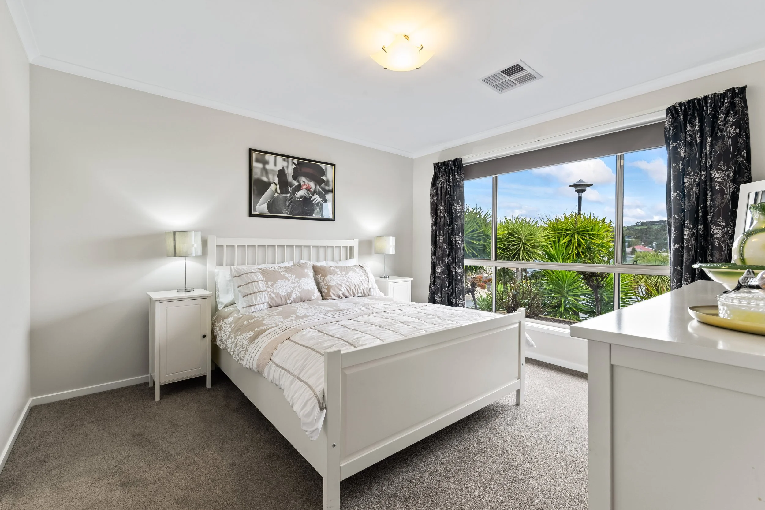 A bright bedroom with a white wooden bed, beige patterned bedding, two matching white bedside tables with lamps, large window with black floral curtains showing greenery outside, wall art of a woman in black and white, and a ceiling light.