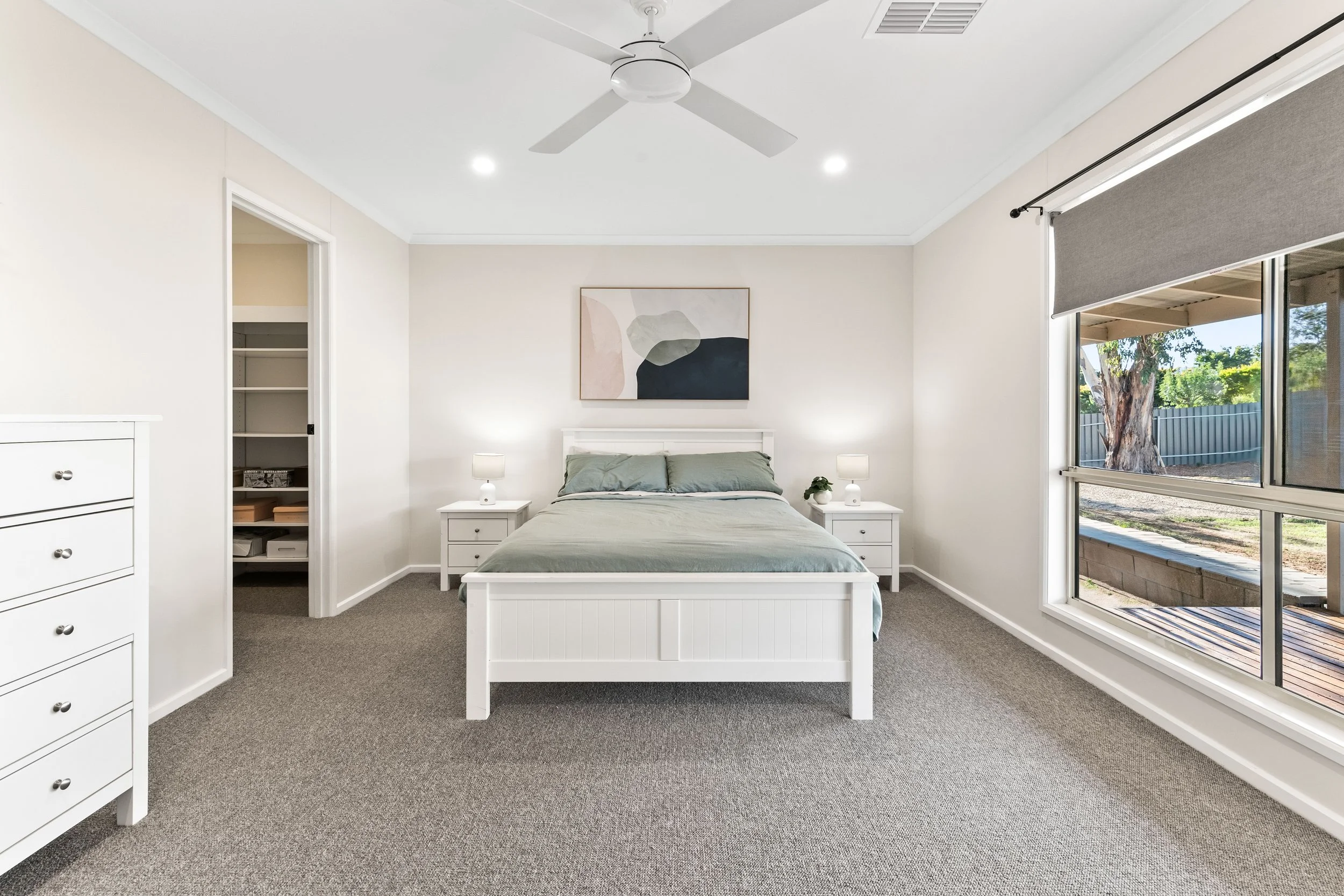 A minimalistic bedroom with a white bed, two nightstands with lamps, a door to a closet, a large window showing trees outside, and a ceiling fan.