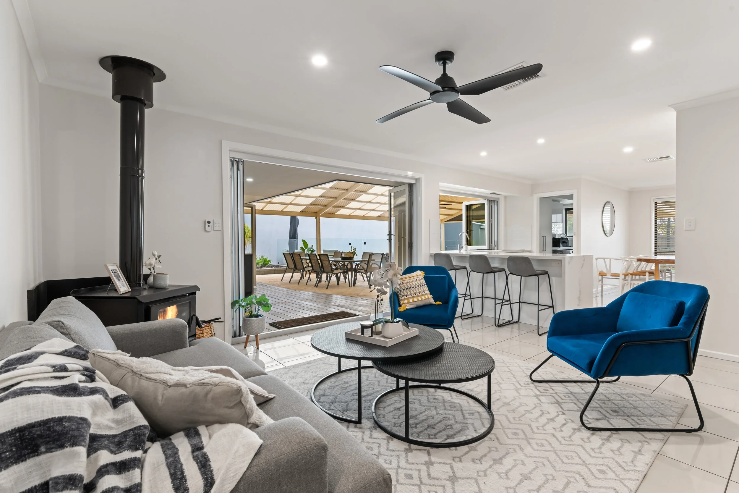 Open-concept living room with a gray sofa, blue armchairs, coffee tables, and a fireplace, with a view of an outdoor patio through sliding glass doors.