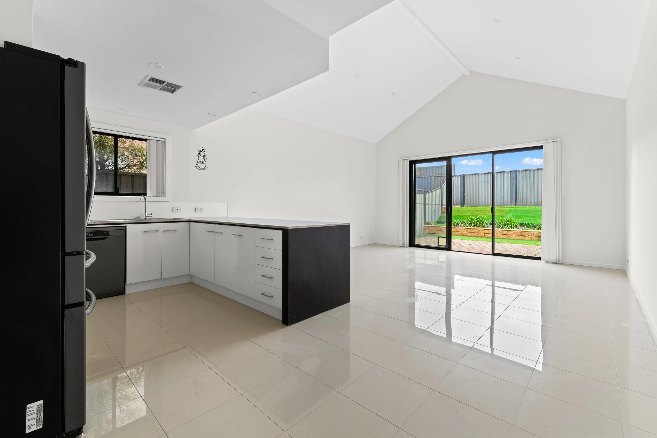 Modern kitchen with white cabinets, black refrigerator, tiled floor, large sliding glass door leading outside to a backyard with grass and a fence, bright natural light, and a window above the sink.