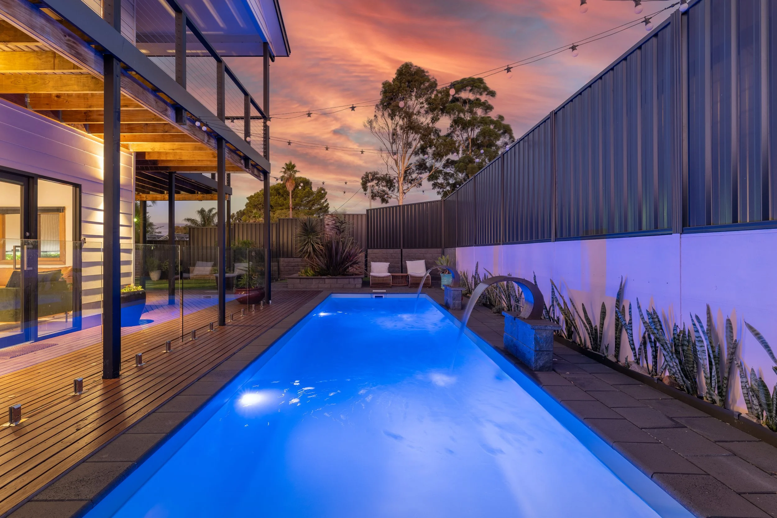 A modern backyard with a swimming pool illuminated by blue lights, a wooden deck, and an outdoor seating area. There are tall trees, string lights, and a sunset sky with orange and pink hues.