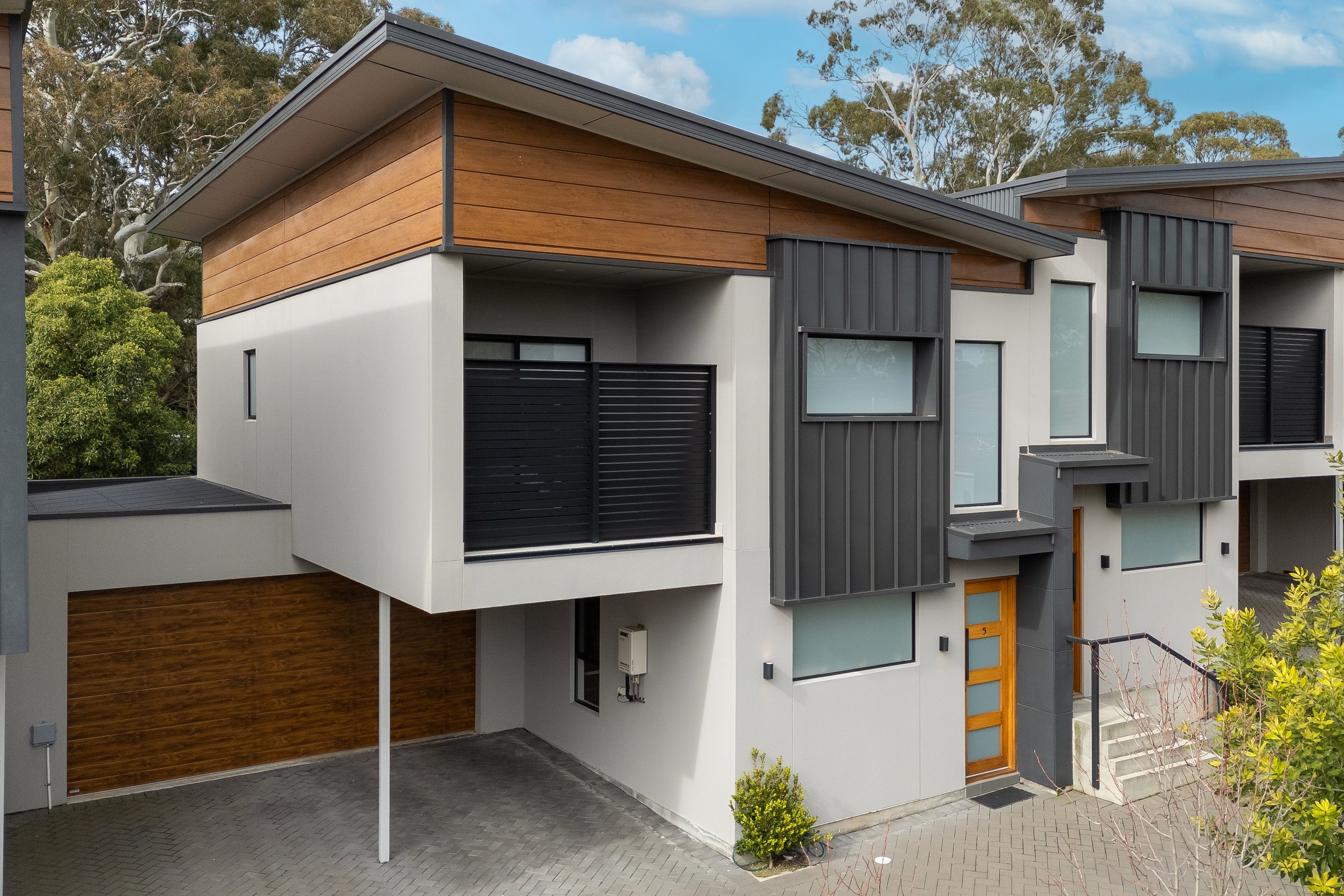 Modern multi-story house with a flat roof, white and black exterior, wood panel accents, multiple windows, and a garage, surrounded by trees and a paved driveway.