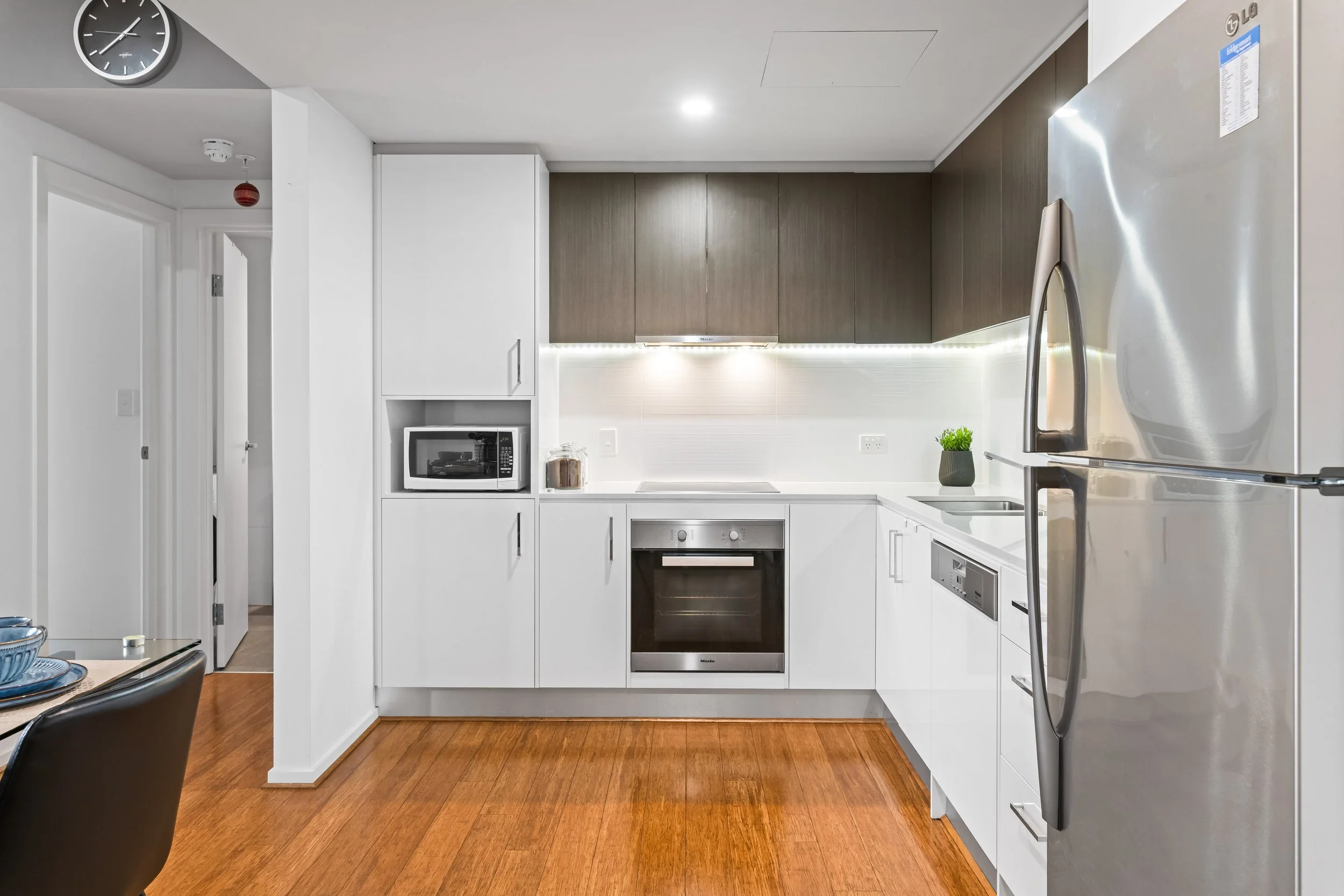 Modern kitchen with white cabinets, stainless steel appliances, brown upper cabinets, a plant on the counter, a microwave, and a clock on the wall.