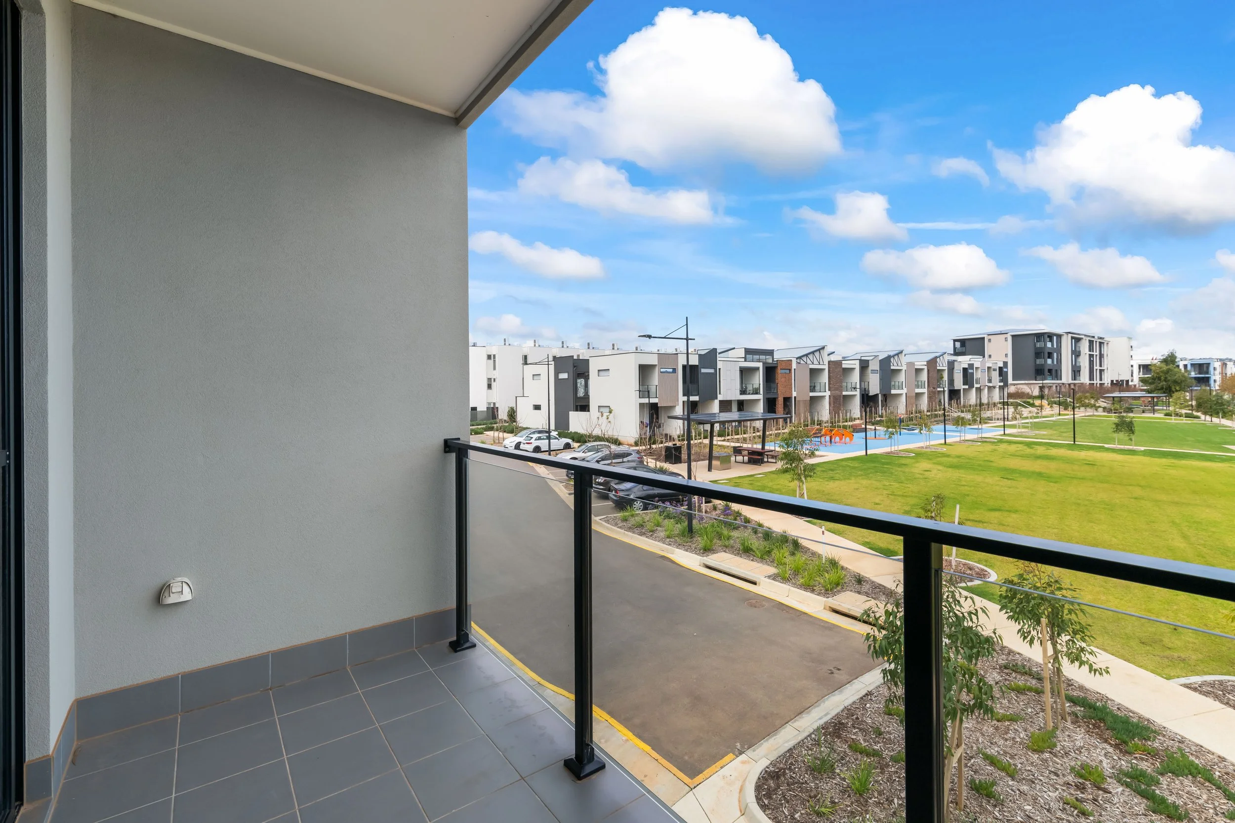 View from a balcony overlooking a modern residential complex with landscaped green lawns, pathways, and a playground, under a partly cloudy sky.