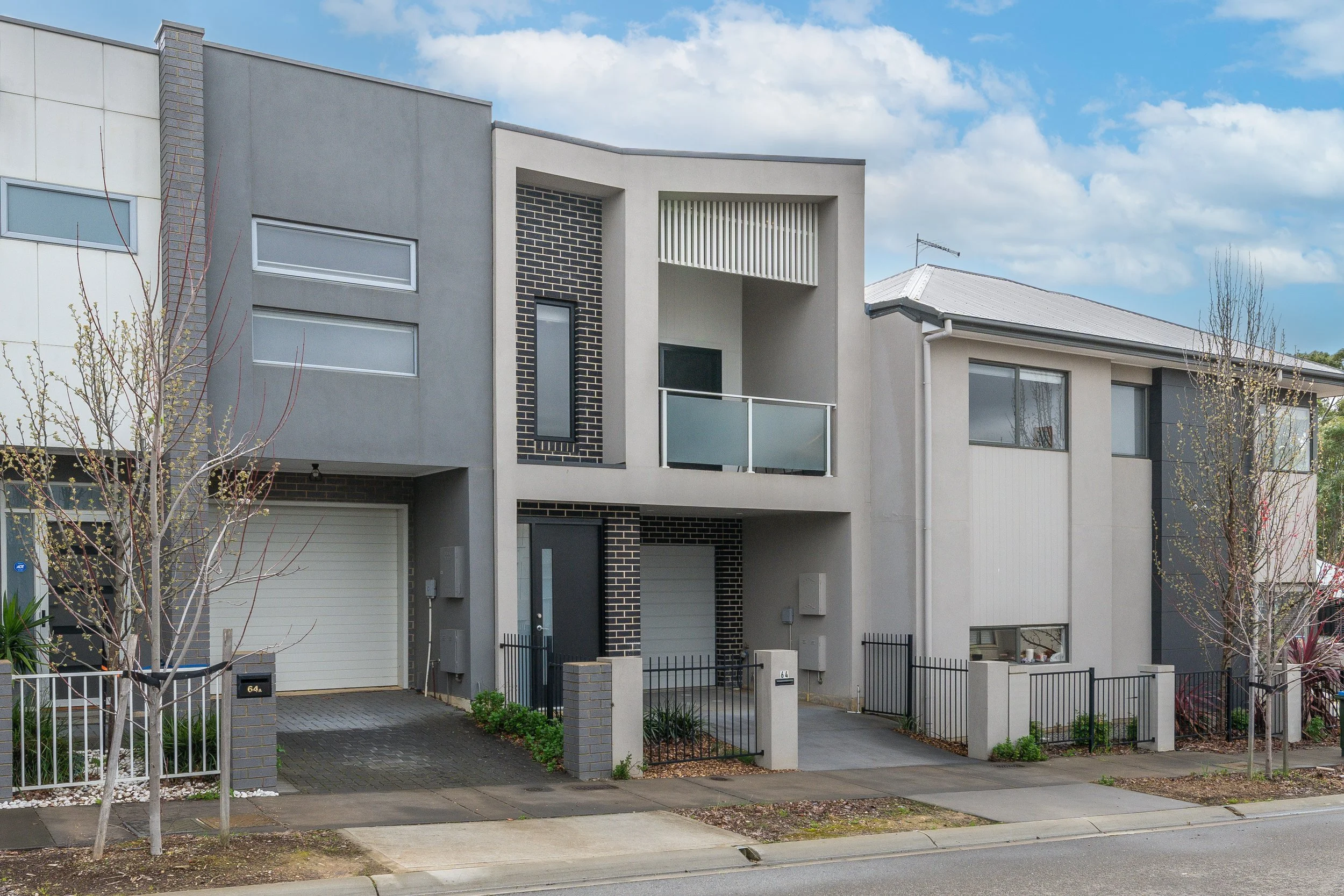 Modern multi-story residential houses with garages, balconies, and fences on a suburban street under a partly cloudy sky.