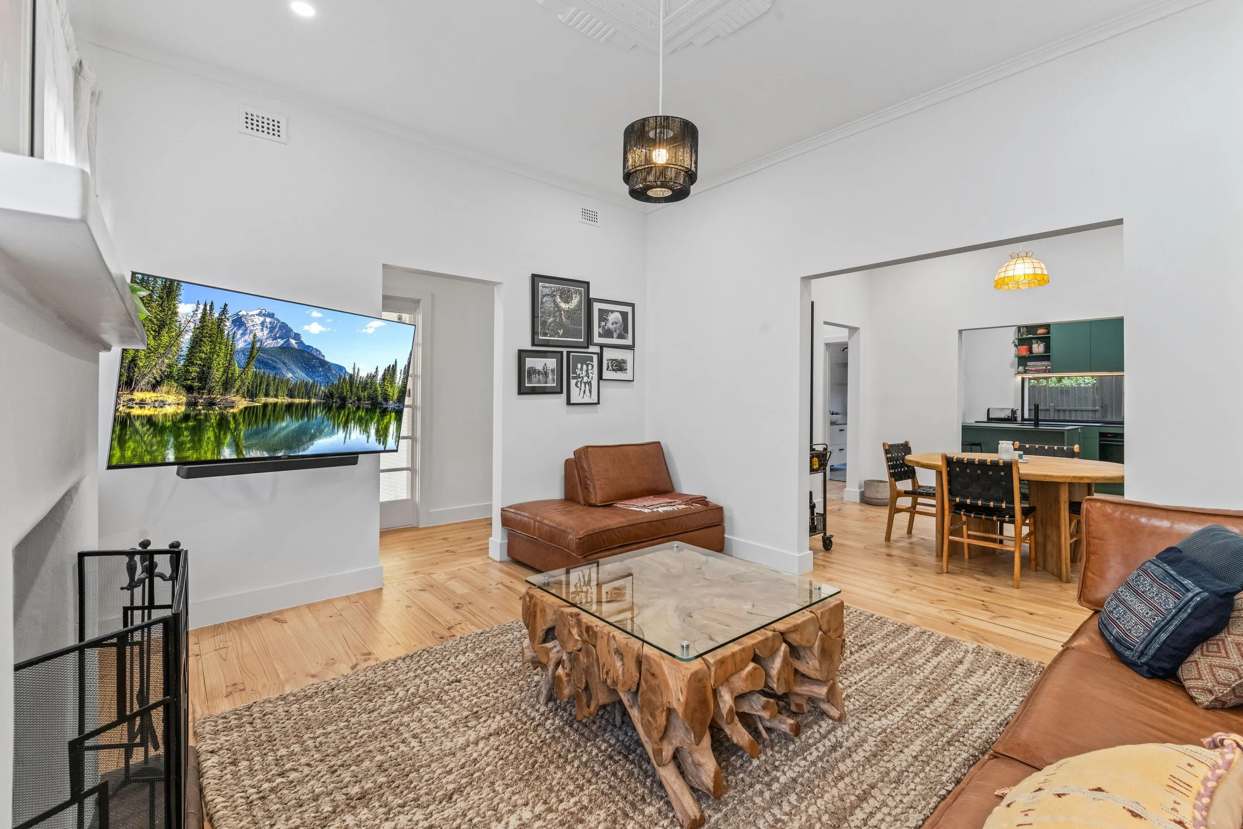 A living room with white walls and ceiling, hardwood flooring, and various furniture including brown leather couches, a glass-topped wooden coffee table, a bench, and a dining table with chairs. A flat-screen TV displays a mountain landscape. Wall ar