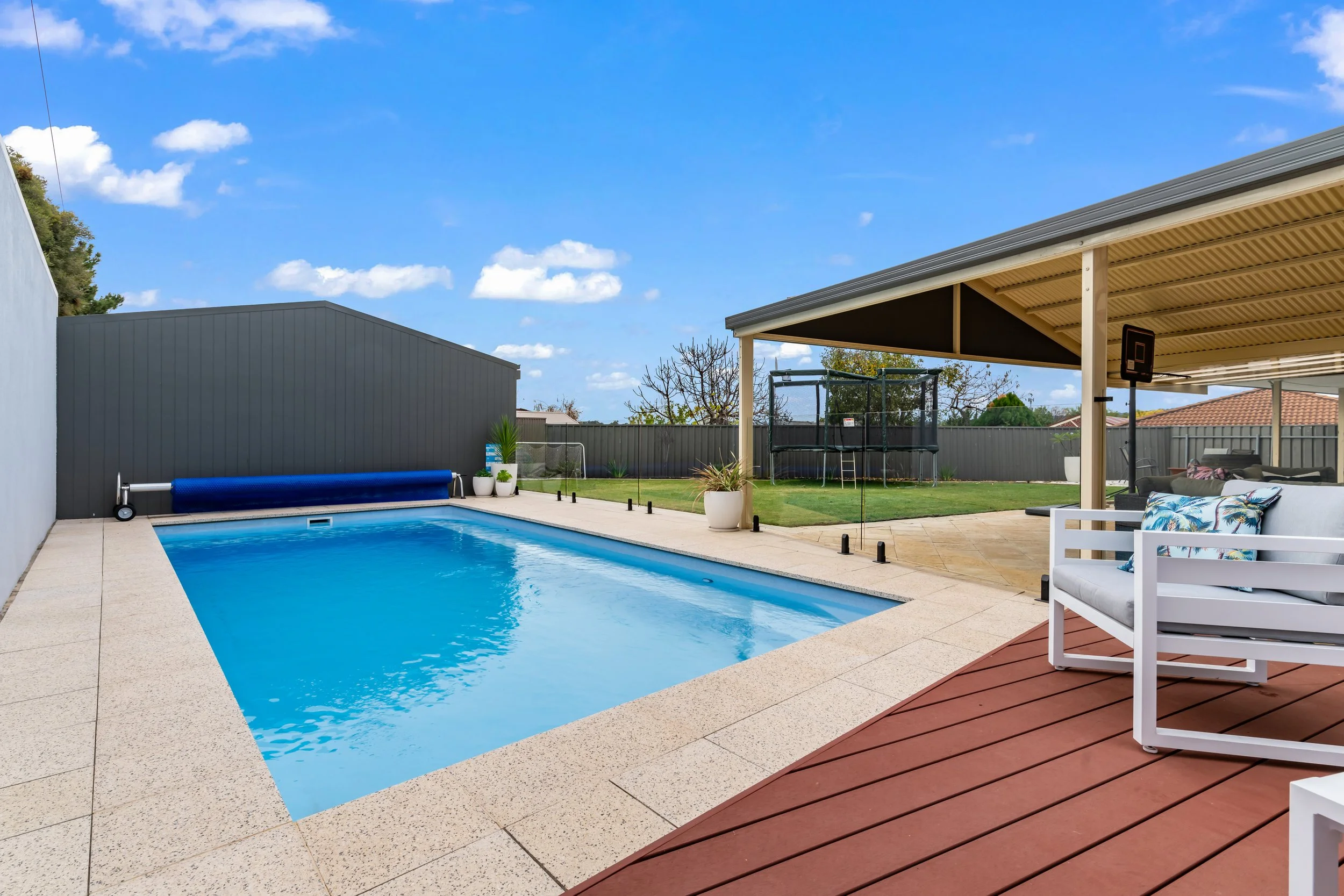 A backyard patio area featuring a swimming pool with a blue safety cover rolled at the edge, surrounded by a beige stone deck, a covered seating area with outdoor furniture, a trampoline, potted plants, and a gray fence under a blue sky with clouds.